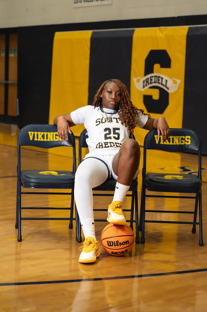 Basketball player in white Vikings uniform poses with ball on court with yellow and black backdrop.