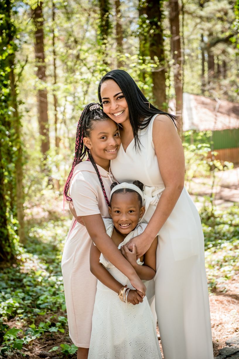 Portrait of a family embracing in sunlit forest setting wearing white and pink dresses.