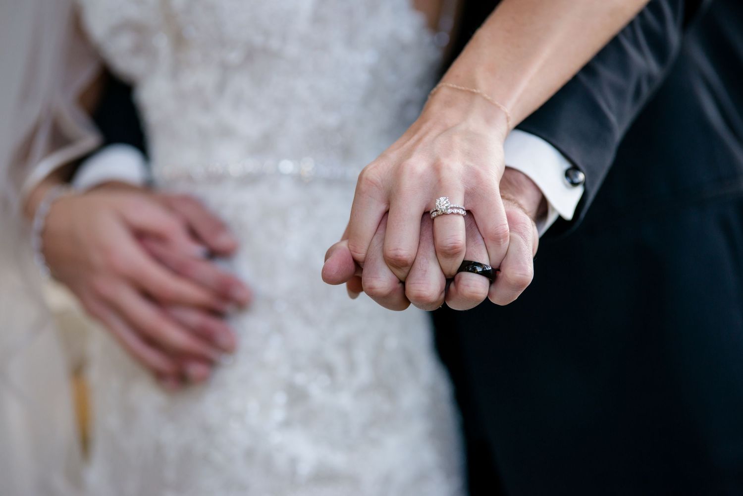 Newlyweds showing off wedding rings, bride in lace gown and groom in black suit, holding hands together.