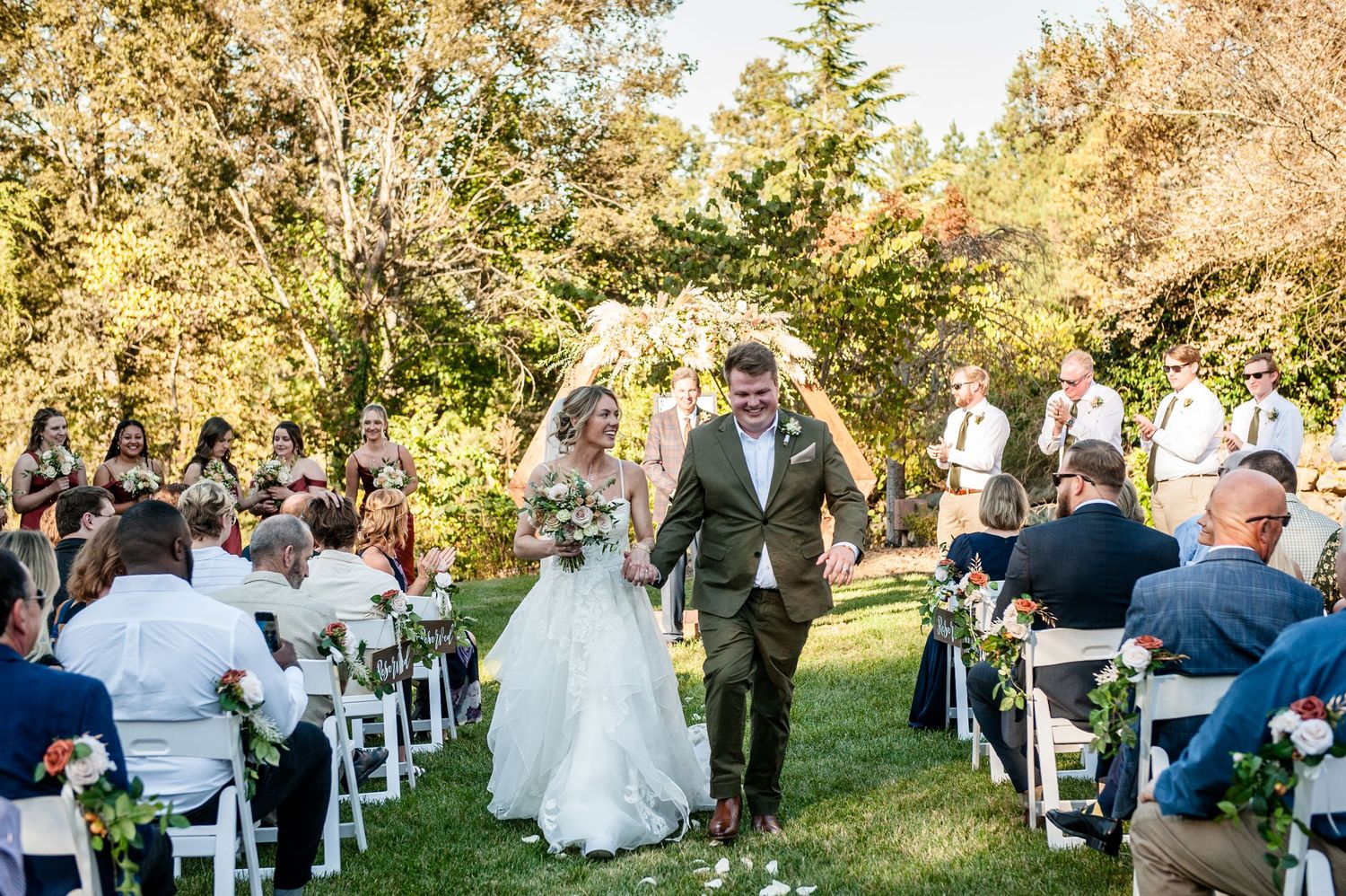 Bride and groom walk down the aisle at an outdoor fall wedding ceremony surrounded by guests and colorful trees.