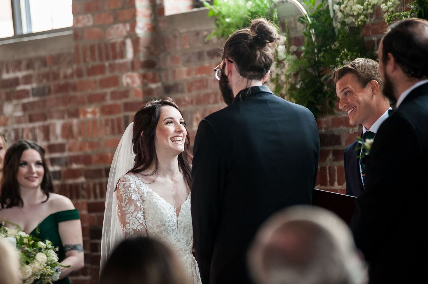 Bride laughing joyfully at wedding ceremony with groom and officiant at brick wall venue.