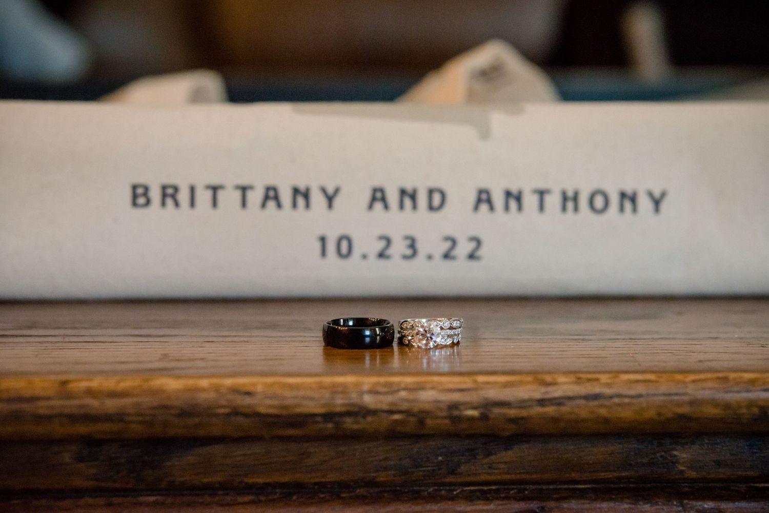 Two wedding rings rest on a wooden surface in front of a personalized bag reading Brittany and Anthony 10.23.22.