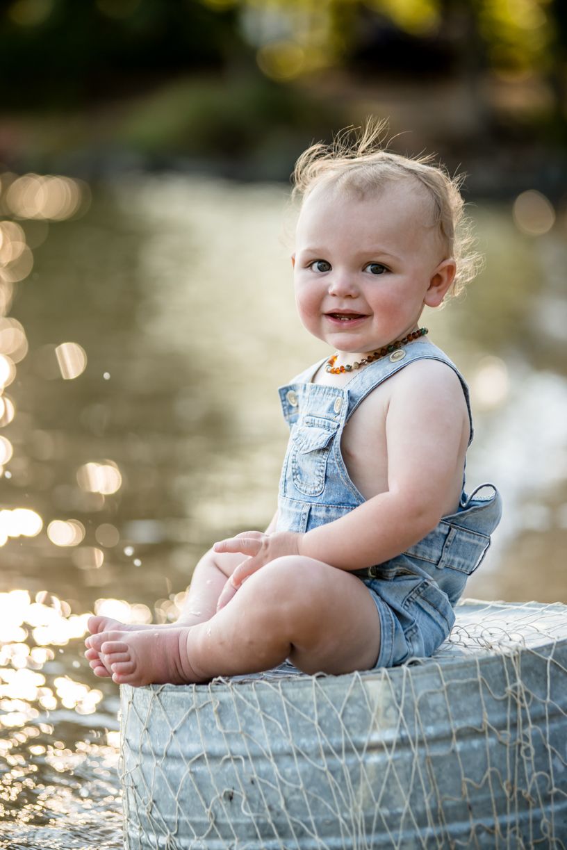 A baby wearing denim overalls sits on a rustic basket outdoors with sparkling water in the background.
