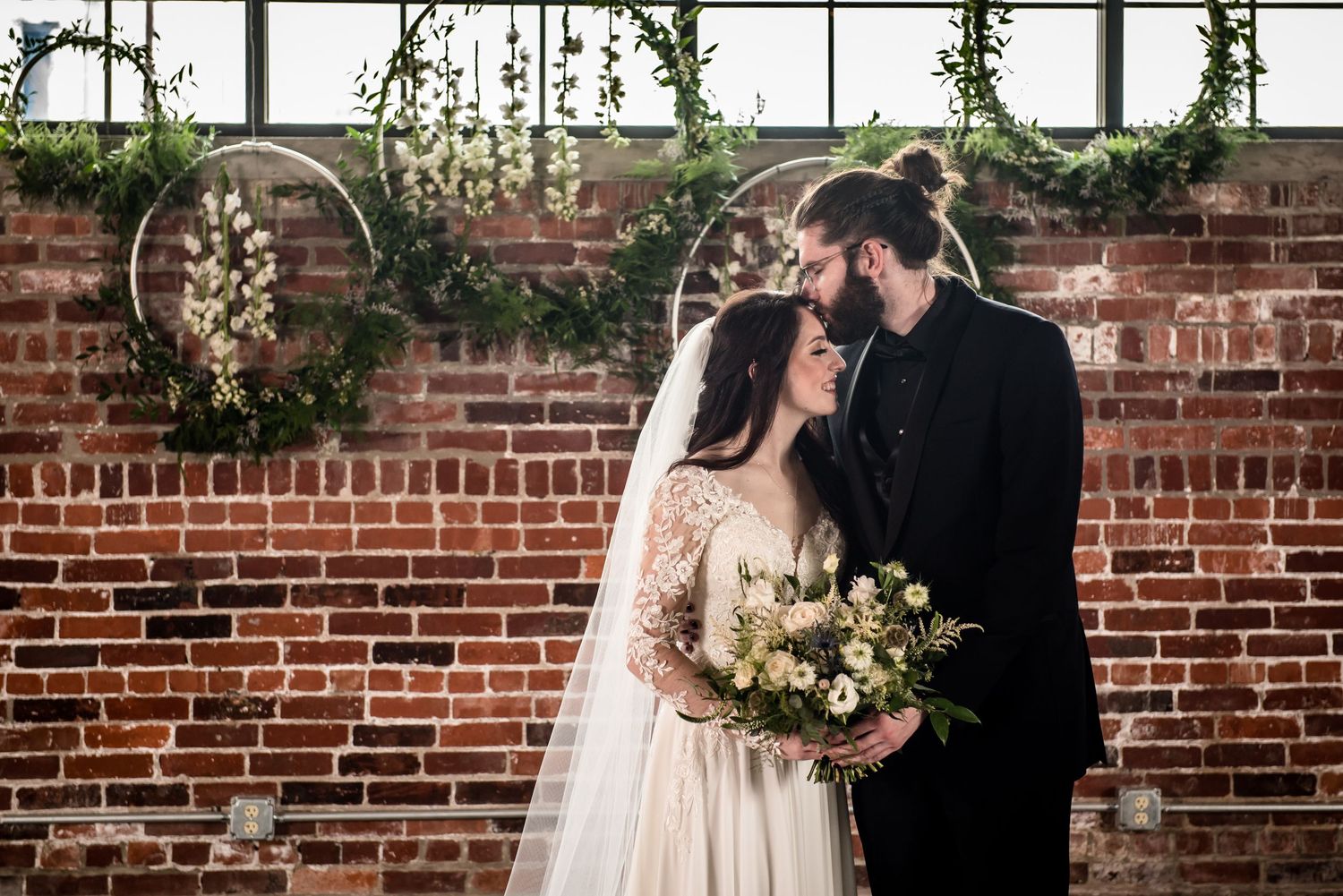 Bride and groom share romantic moment in front of rustic brick wall with circular floral hoops at industrial wedding venue.