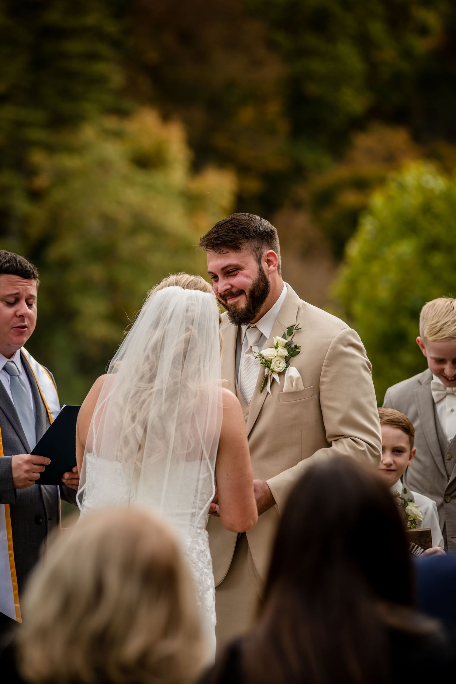 Groom in tan suit smiling joyfully at bride during outdoor wedding ceremony with lush green trees in background.