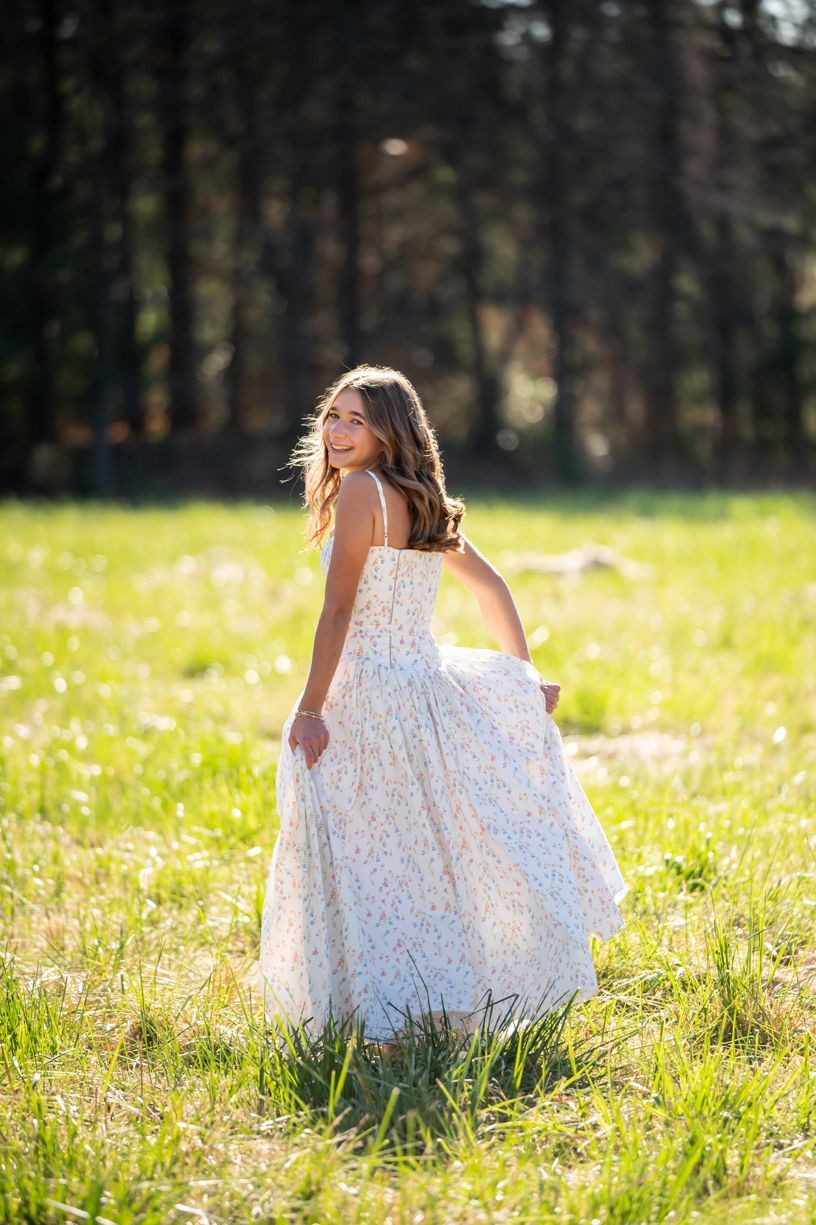 A senior Girl in white dress runs through sunny meadow with wildflowers.