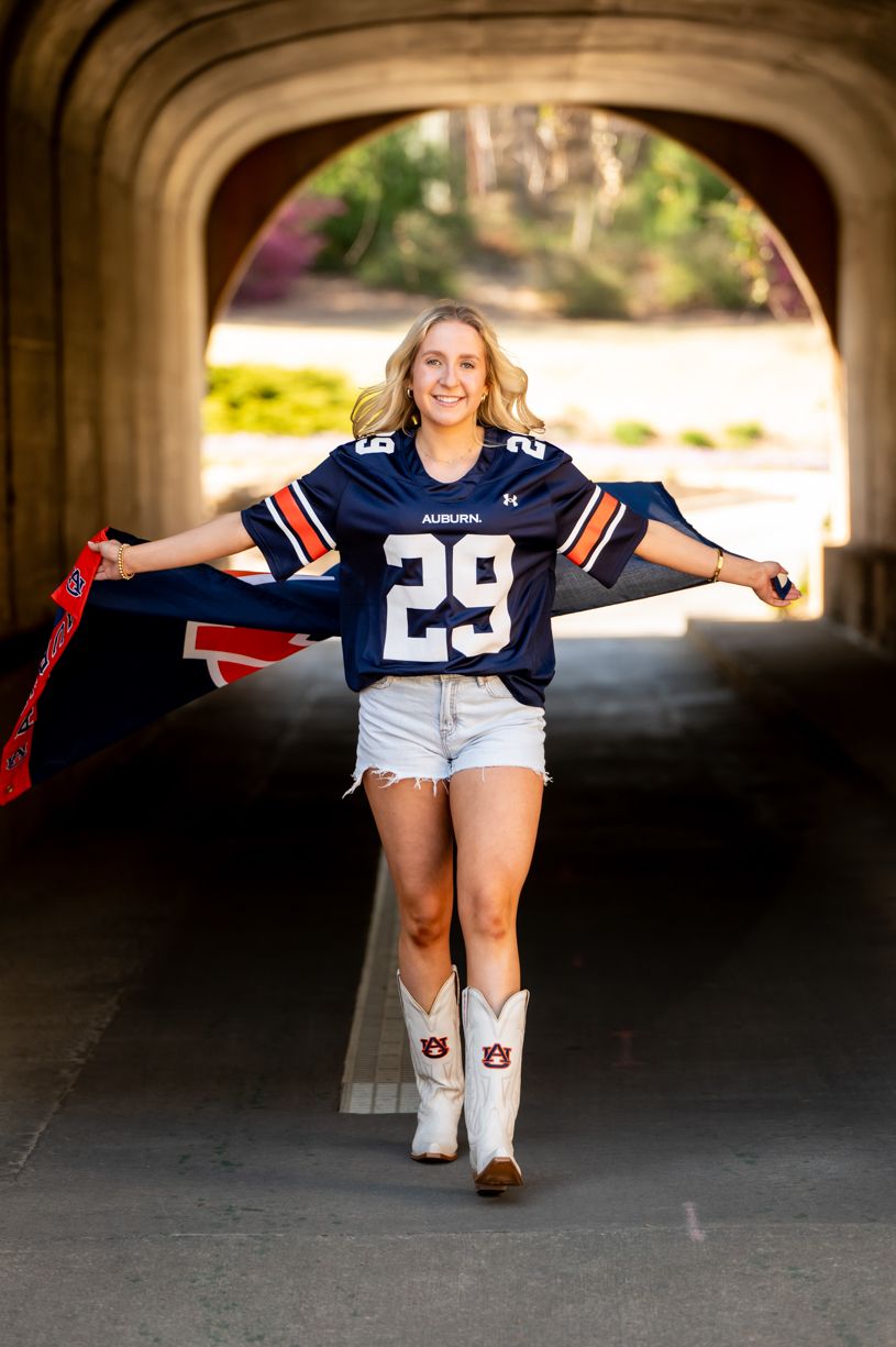Senior girl poses in navy jersey and denim shorts under concrete archway.