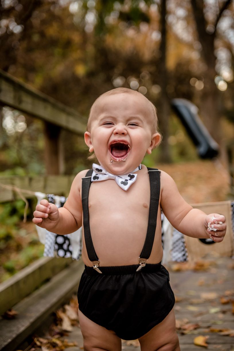 Happy baby wearing bow tie and suspenders laughs while standing on a wooden bridge.
