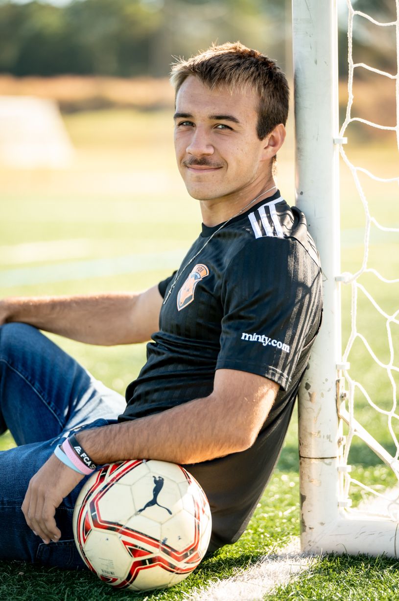A senior soccer player relaxes against a goal post with a ball.