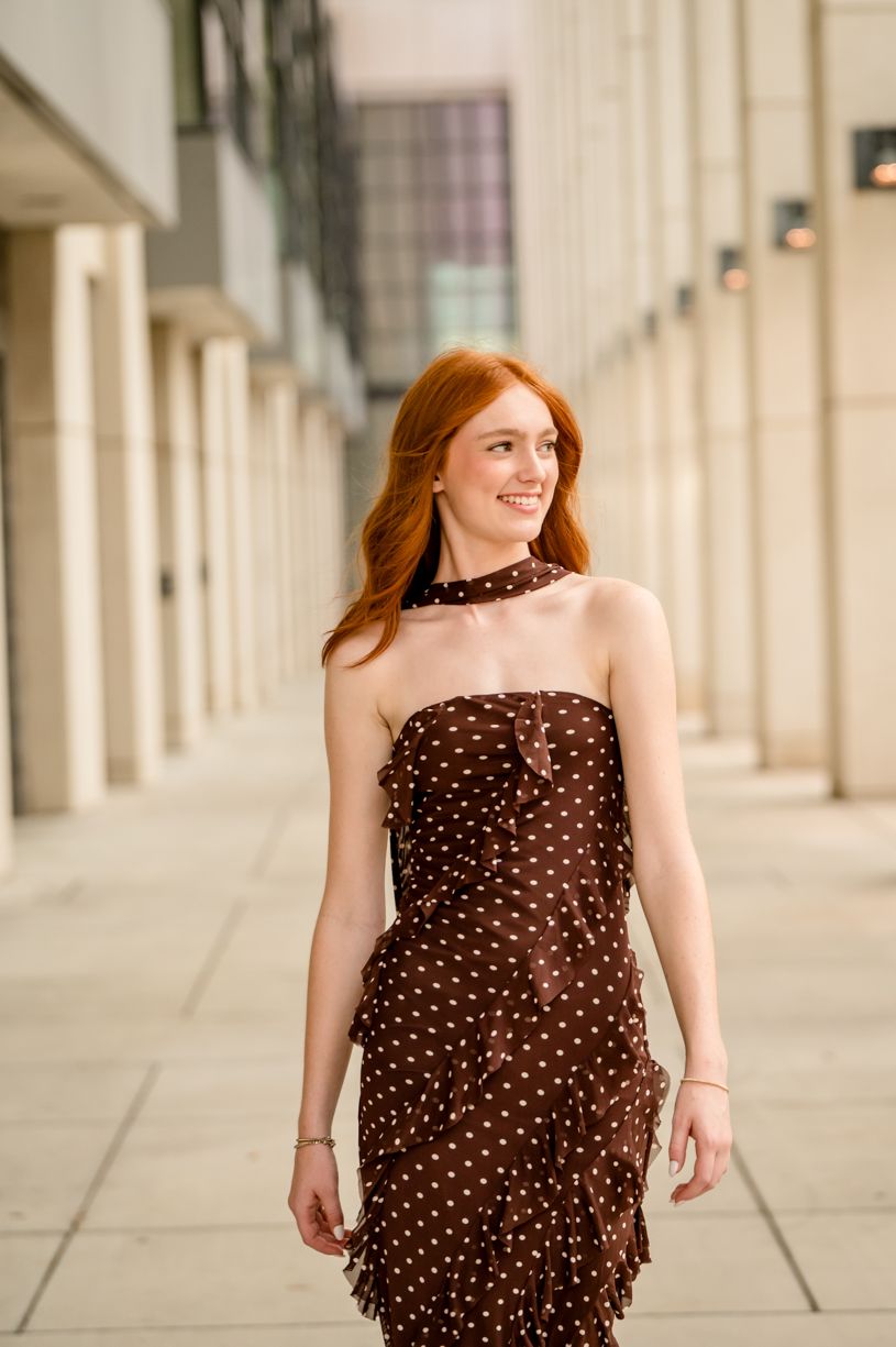 Urban portrait of a senior girl wearing a brown polka dot dress against architectural columns.