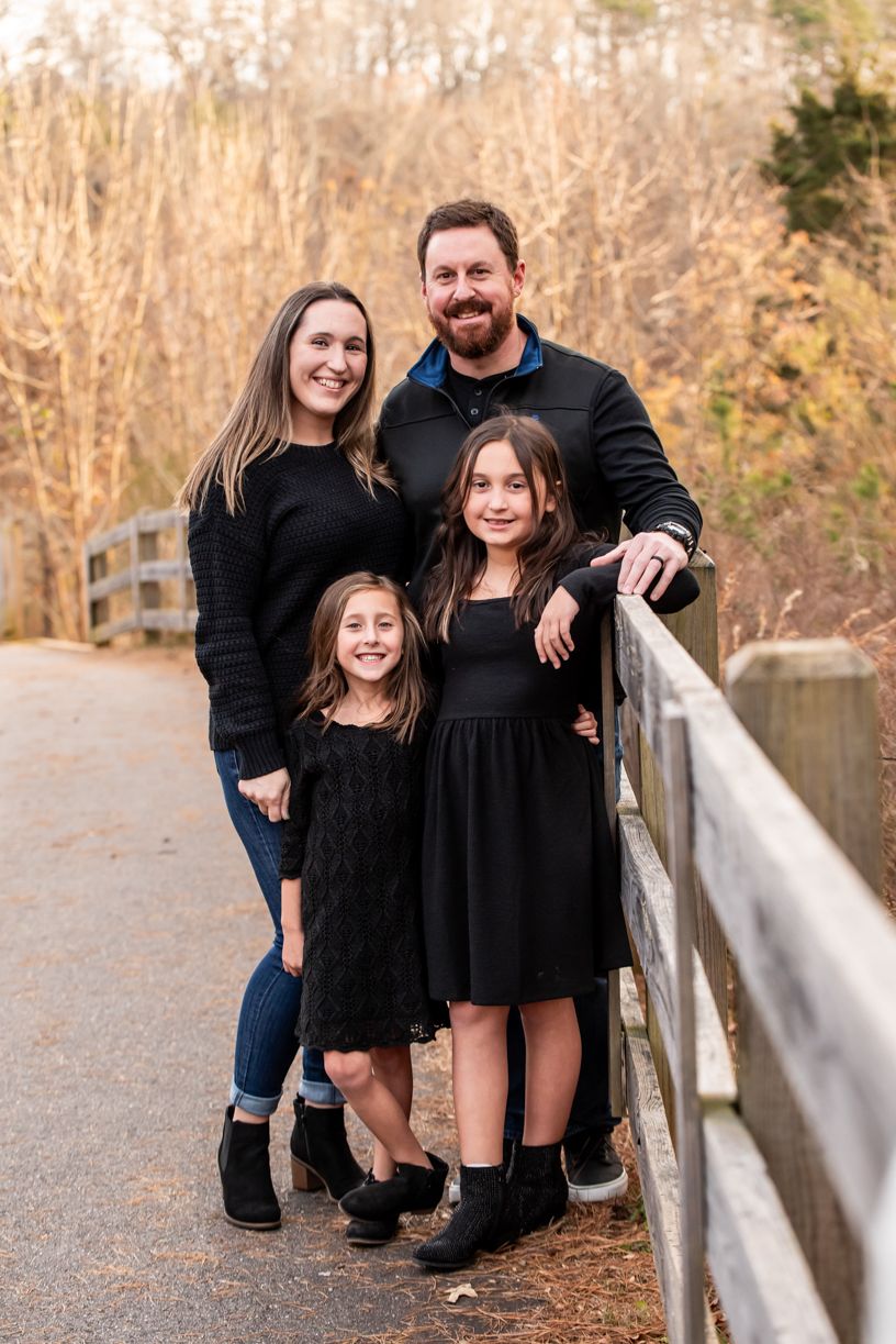 A family of four wearing coordinated black outfits poses together on a wooden bridge.