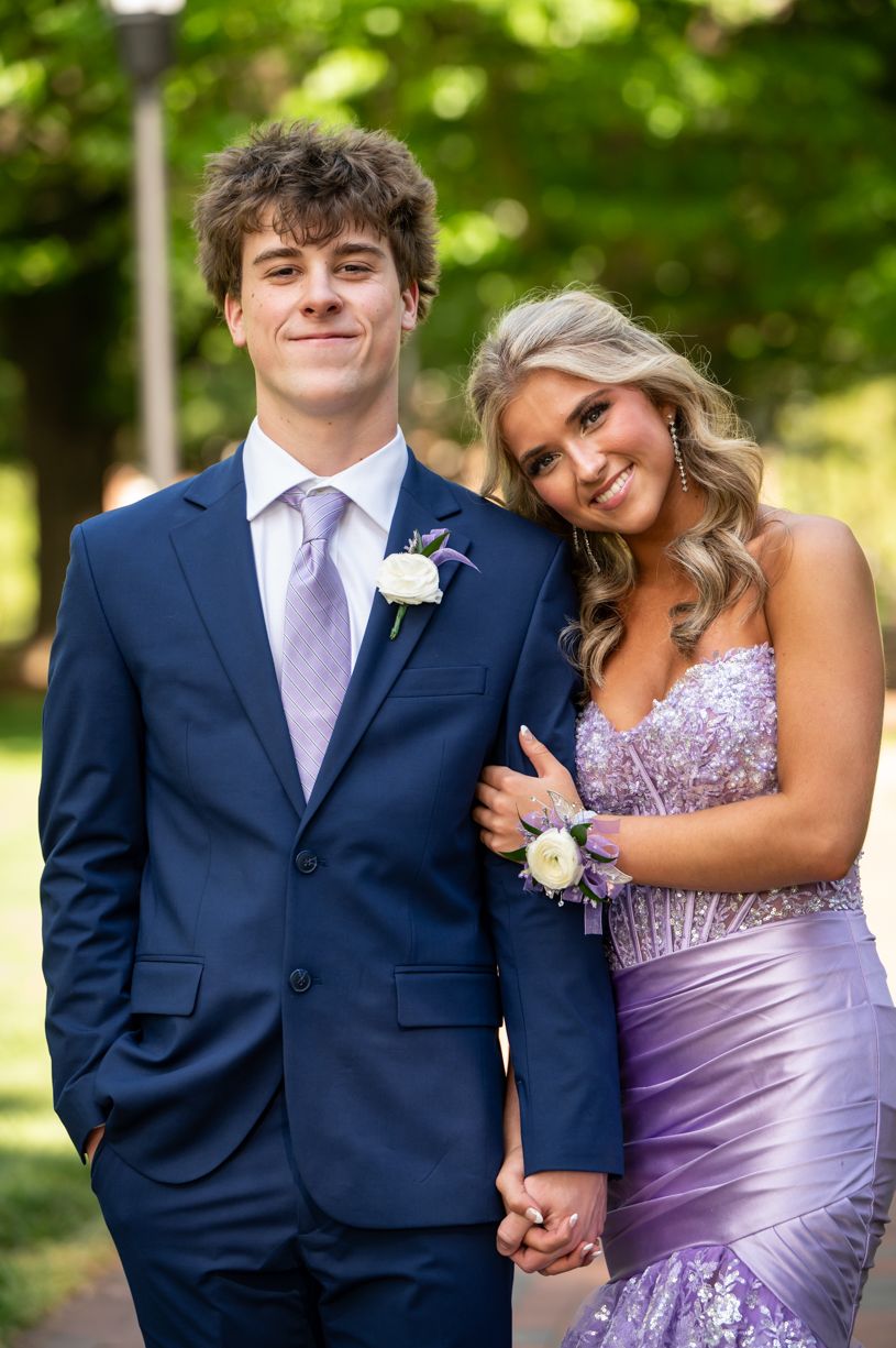 Formal prom couple in navy suit and purple dress posing outdoors with corsage and boutonniere.