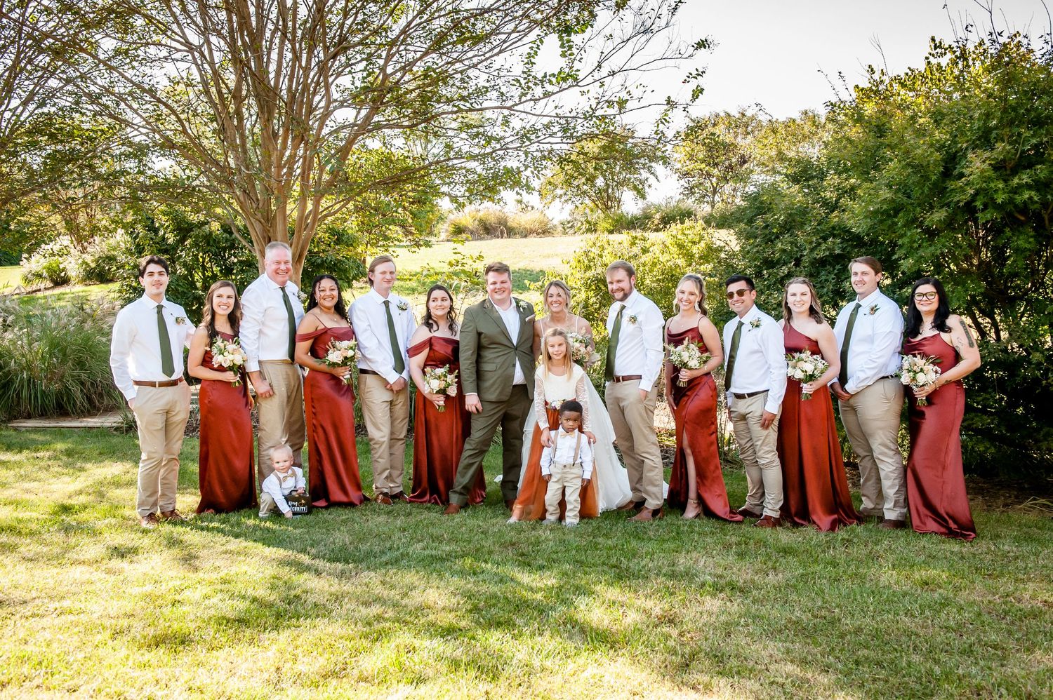 Wedding party in red dresses and tan suits posing outdoors on green lawn with trees in background.