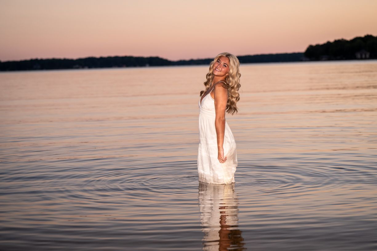 A senior girl Sunset photos at a lake with white clothing and calm water reflections.