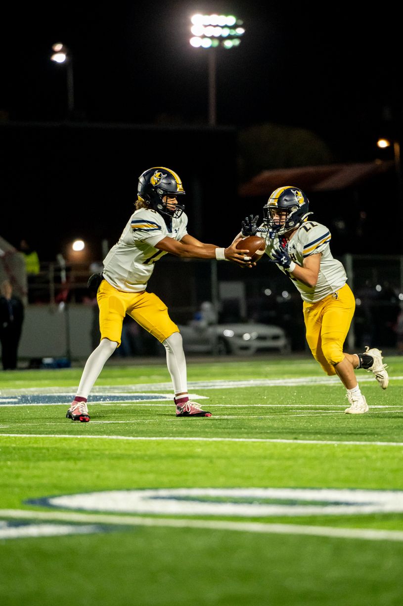 Football players in yellow uniforms executing handoff play under stadium lights.