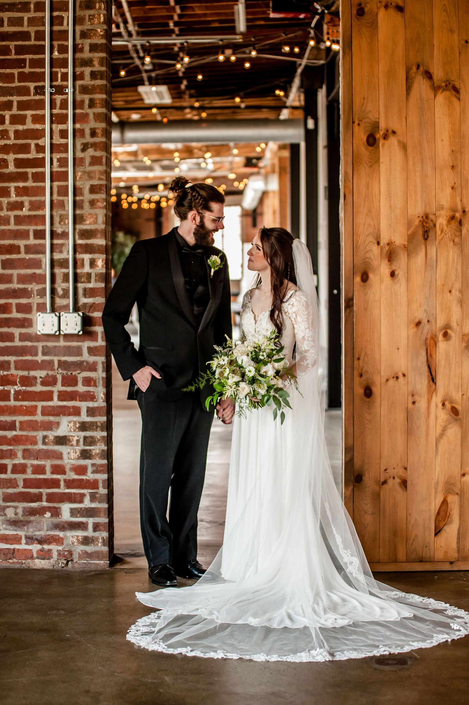 Bride and groom share a romantic moment in a rustic brick venue, she holds a lush bouquet wearing a long veil.