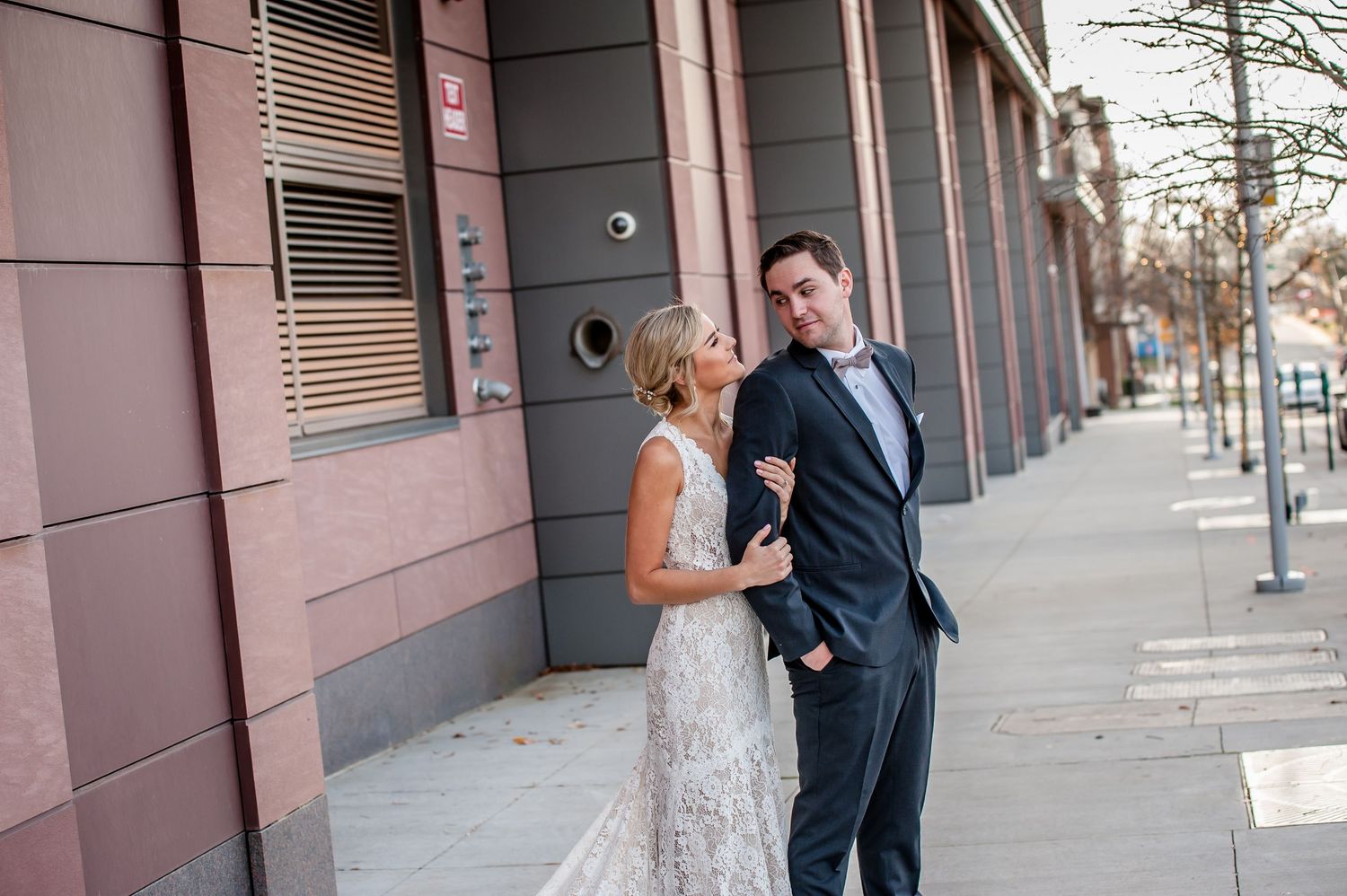 A bride and groom share a romantic moment on a city sidewalk, with the bride in a lace gown and groom in a navy suit.