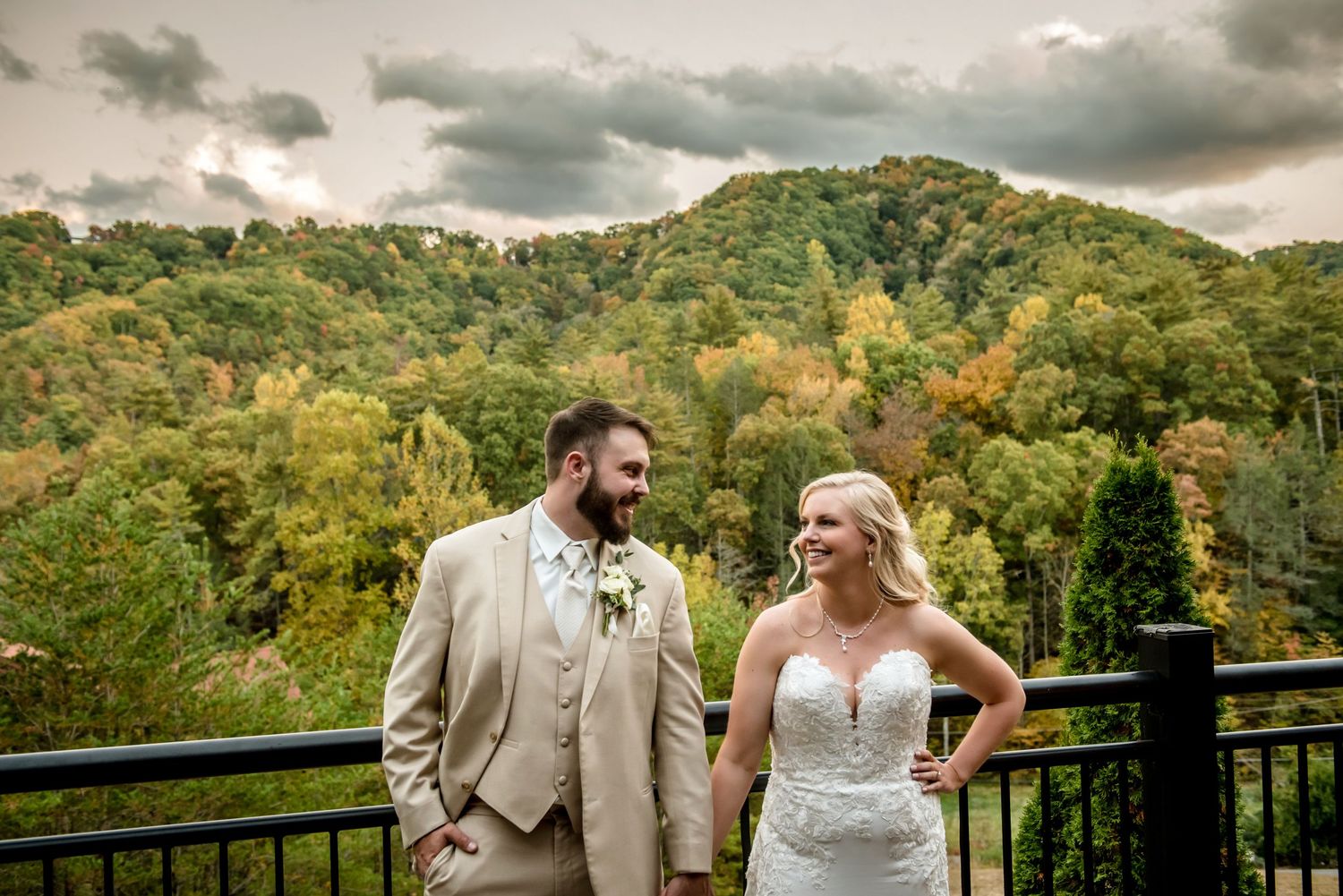Bride and groom share a romantic moment on a deck overlooking lush autumn forest hills under dramatic cloudy skies.