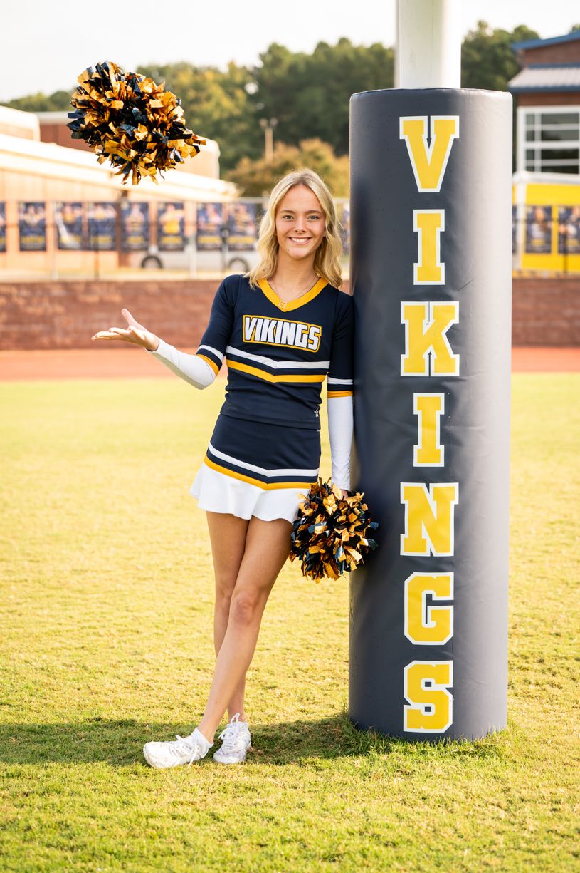 Cheerleader poses with gold and black pom poms on sunny football field with Vikings team branding.