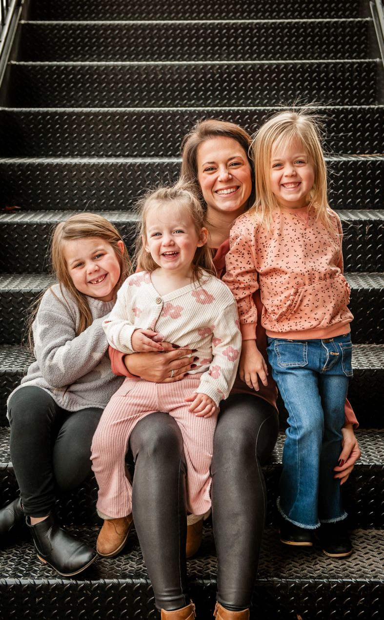 A joyful family sits together on industrial metal stairs wearing coordinated casual outfits in pink and gray tones.