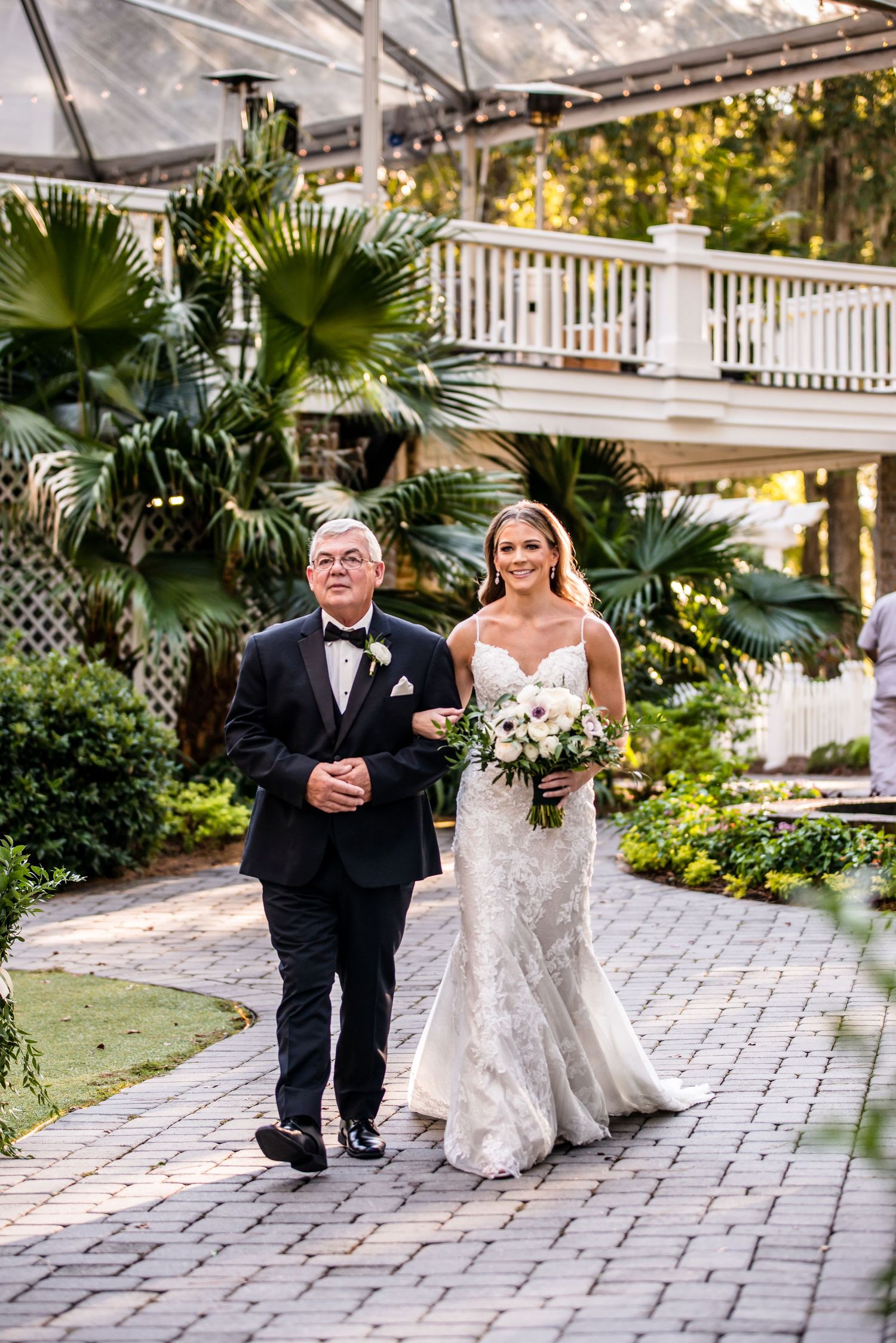 Bride in lace gown walks down brick aisle with escort amid lush tropical greenery at outdoor wedding venue.