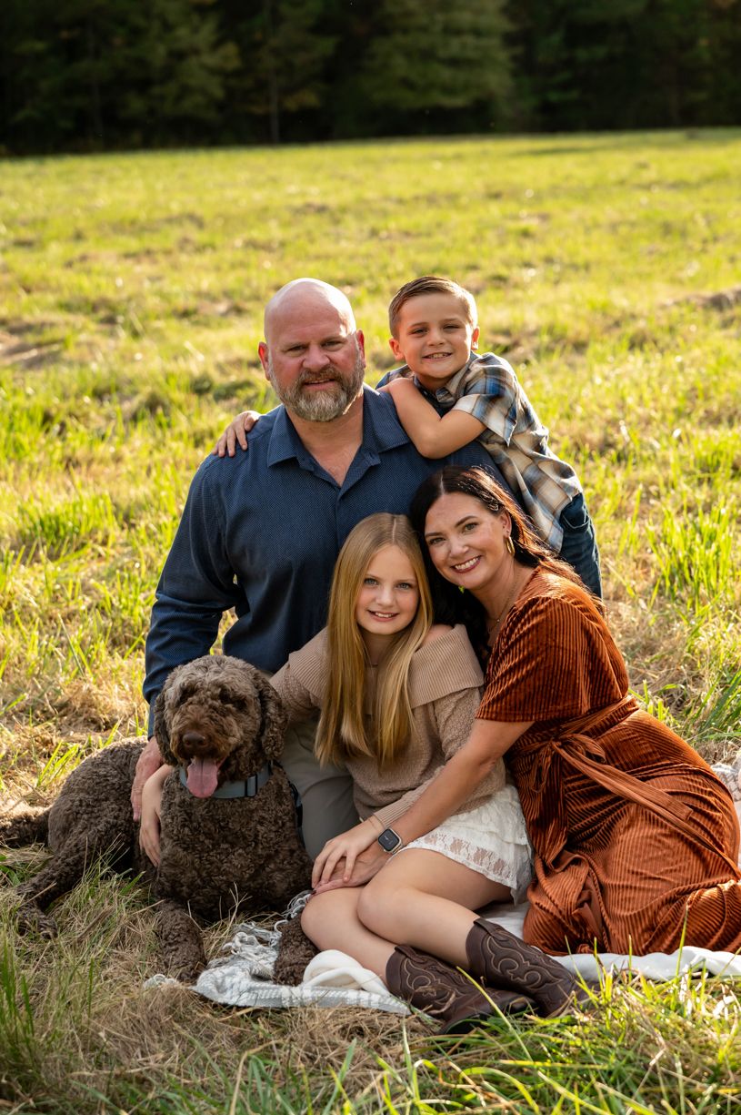 A family sits together in a golden field during sunset with their dog, sharing warm embraces and smiles.