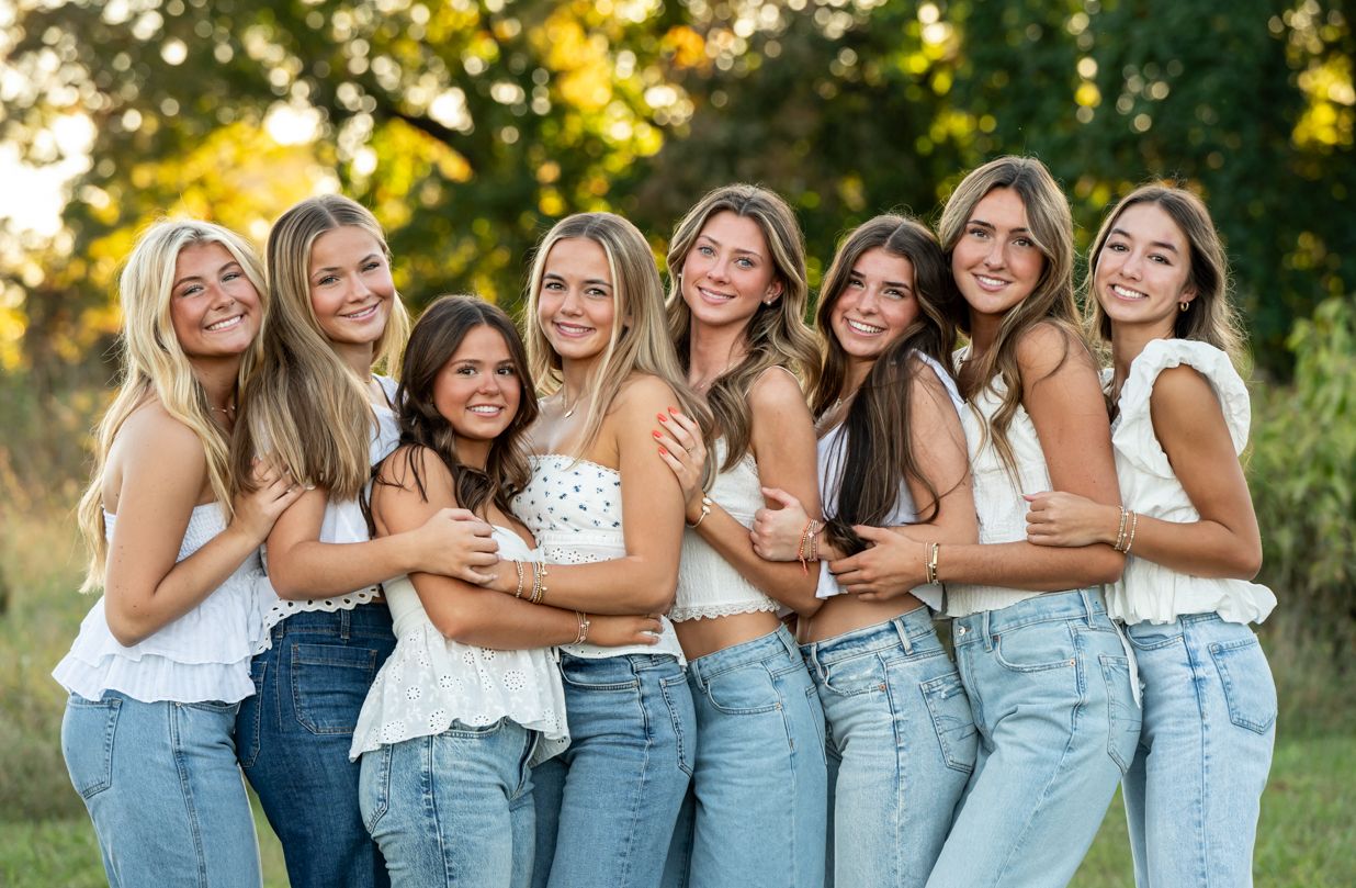 A group of senior girls wearing coordinated denim and white outfits poses together outdoors during a sunny golden hour photoshoot.