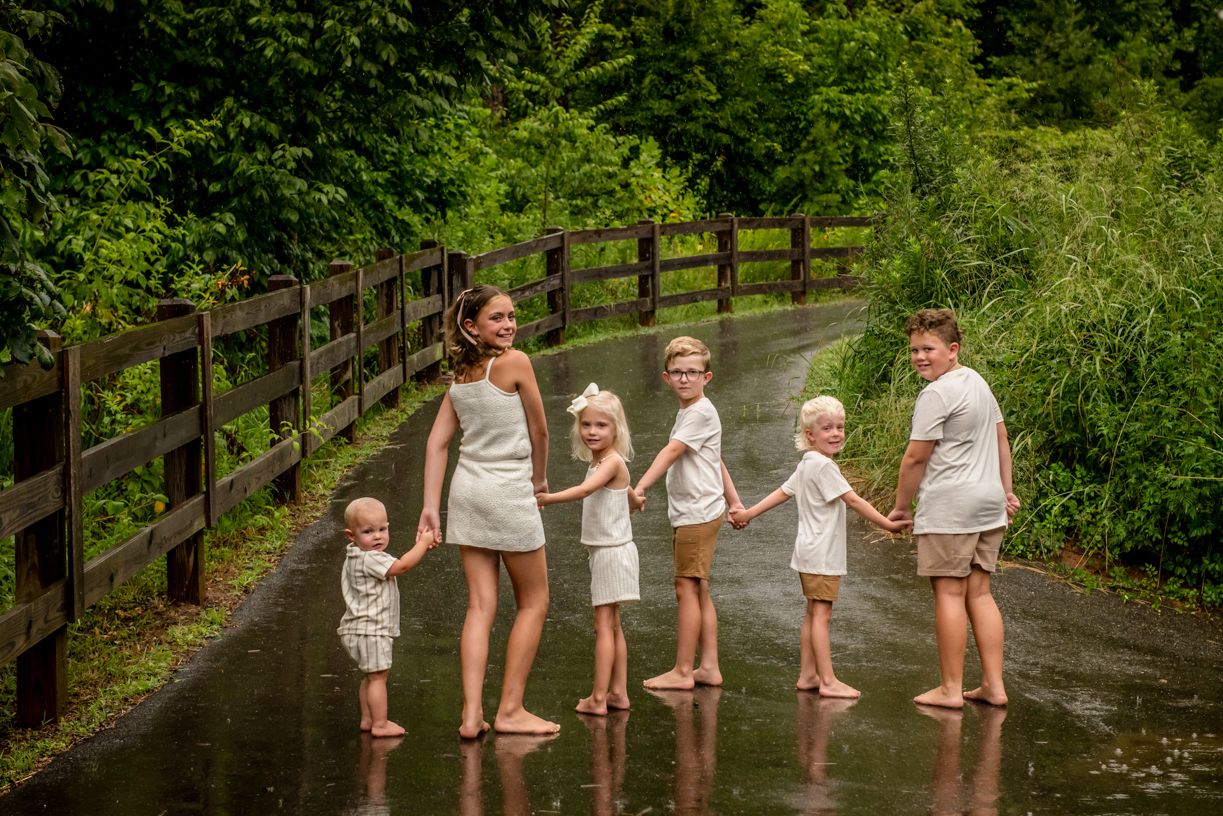 A line of children wearing matching white outfits stands along a wooden fence in a lush green setting.