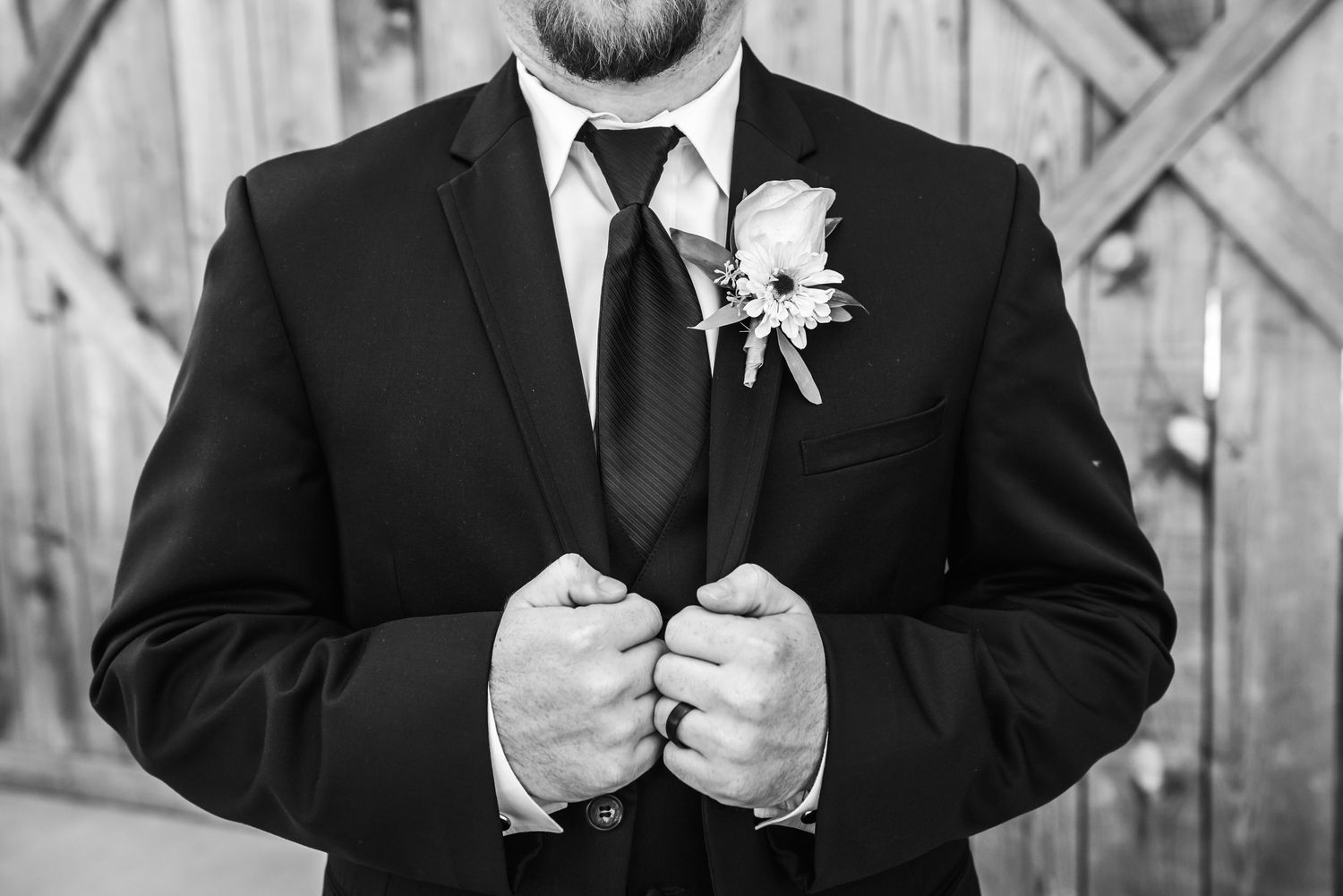 Black and white photo of a groom in a suit adjusting his jacket, wearing a boutonniere and wedding ring.