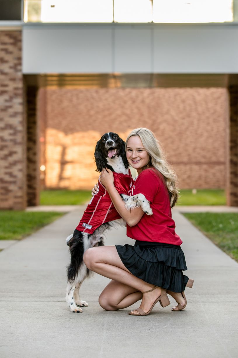 Senior girl in red shirt kneeling next to black and white dog on sidewalk.
