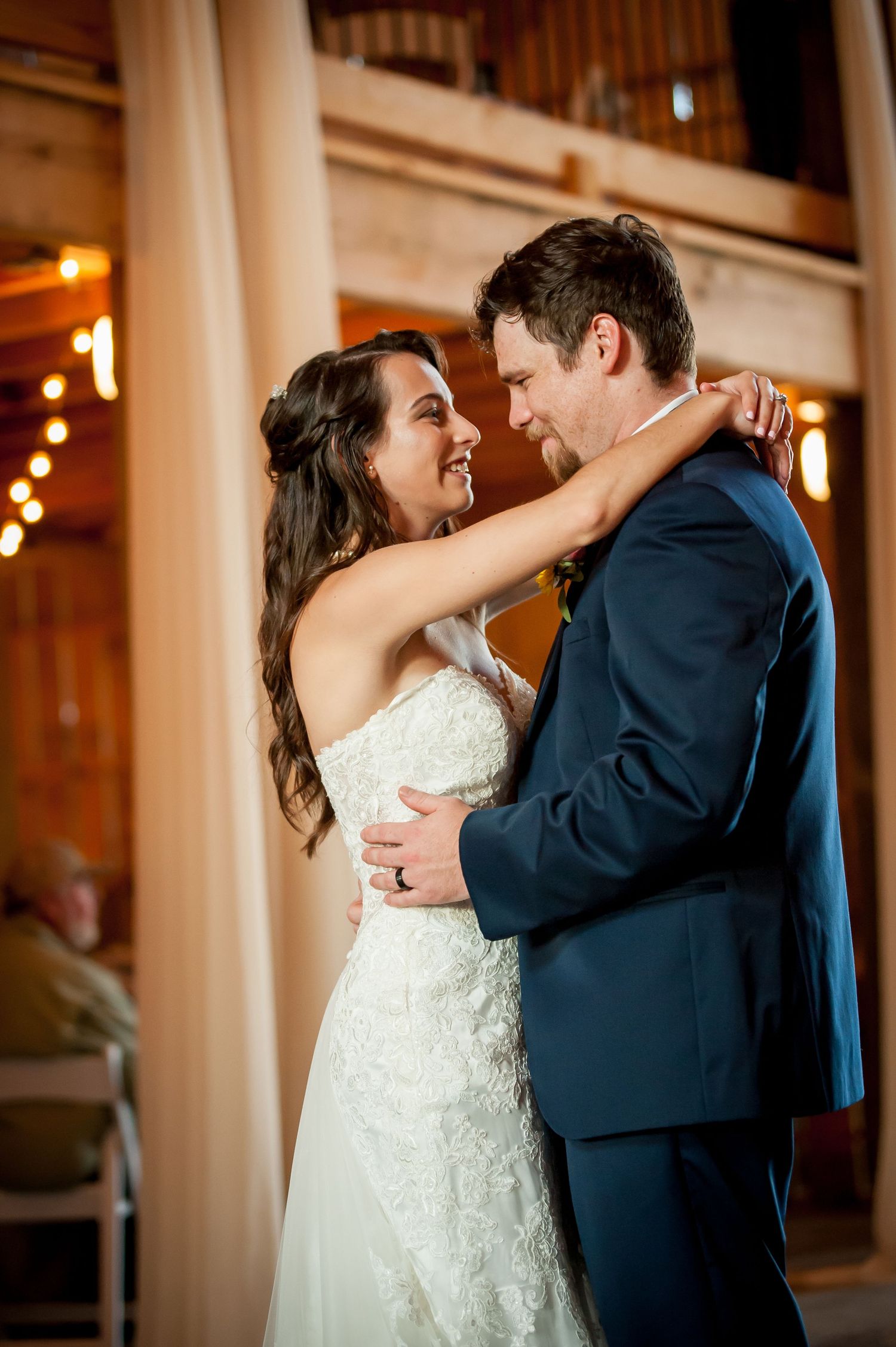 Bride and groom share their first dance in a warmly lit barn venue, gazing lovingly at each other.