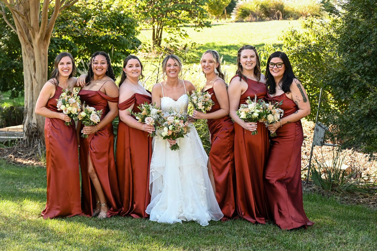 Bride in white gown poses with six bridesmaids in rust-colored satin dresses holding white bouquets outdoors.