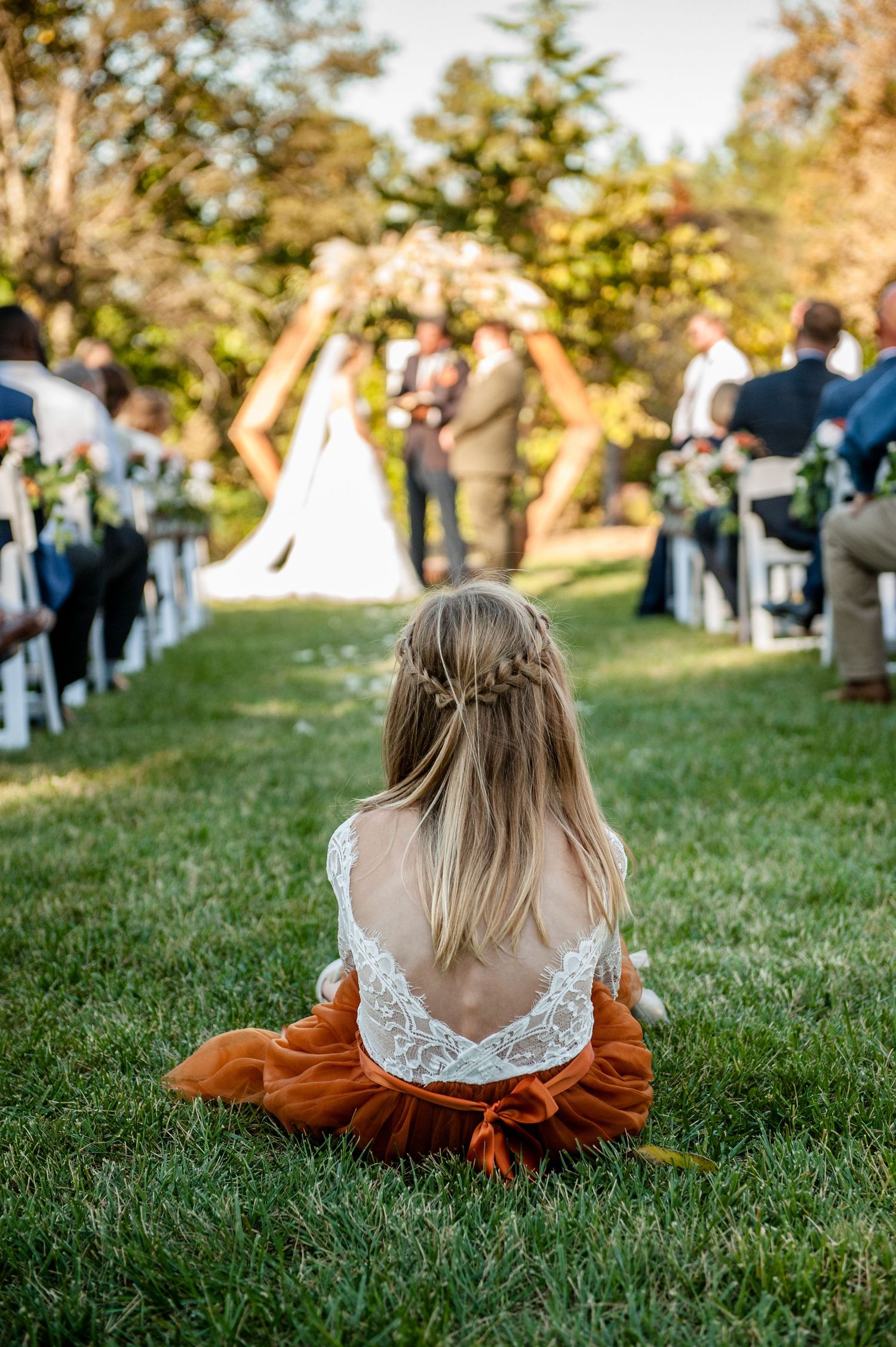 Young flower girl in lace and rust dress sits on grass watching outdoor wedding ceremony with hexagonal arch.