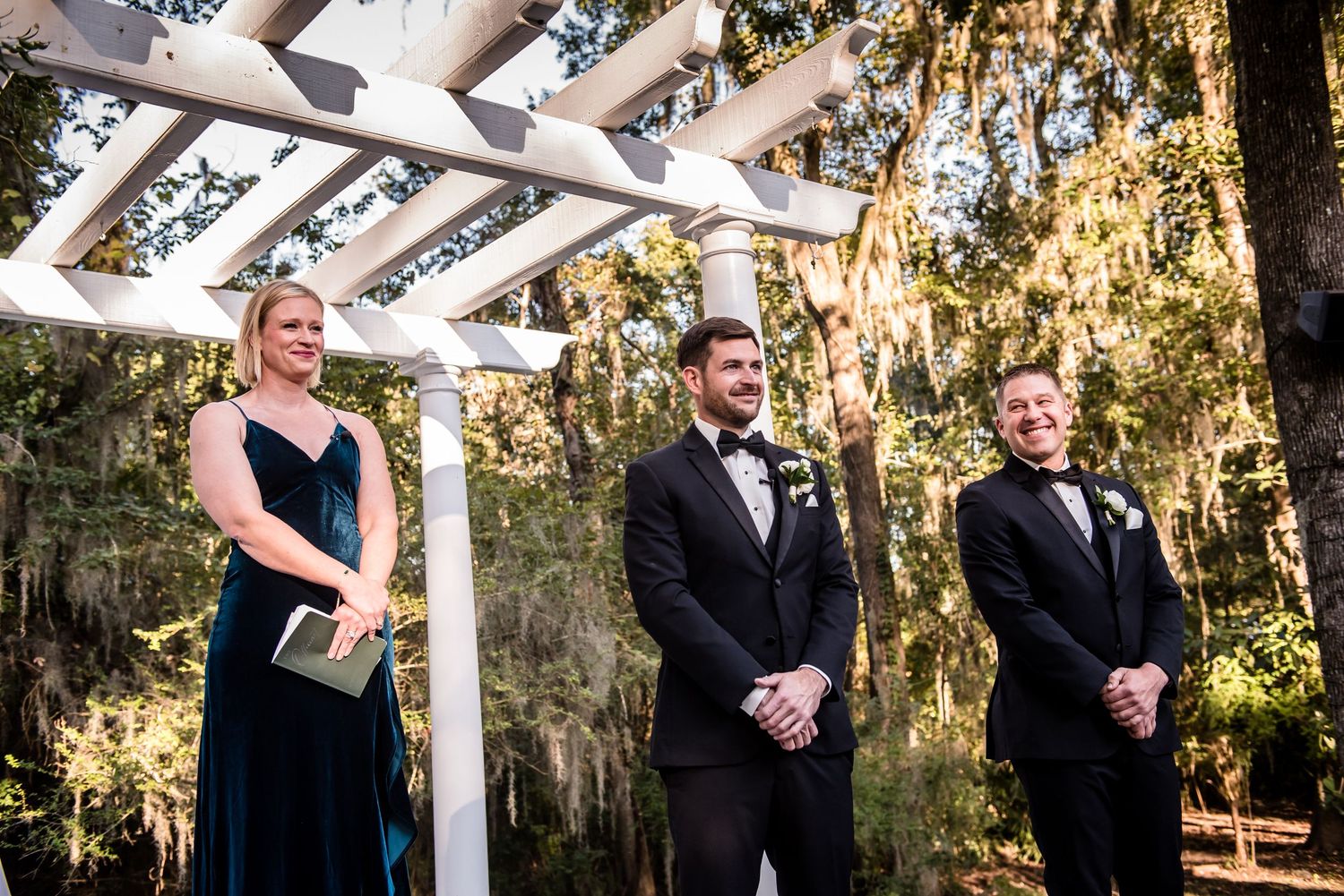 Groom and groomsmen stand under white pergola arbor at outdoor wedding ceremony surrounded by moss-draped trees.