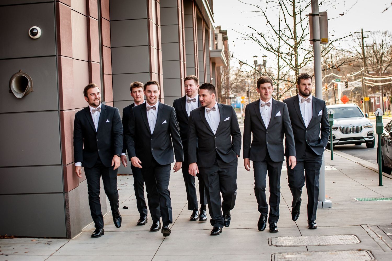 Seven groomsmen in black tuxedos with white bow ties walking confidently down a city sidewalk on a wedding day.
