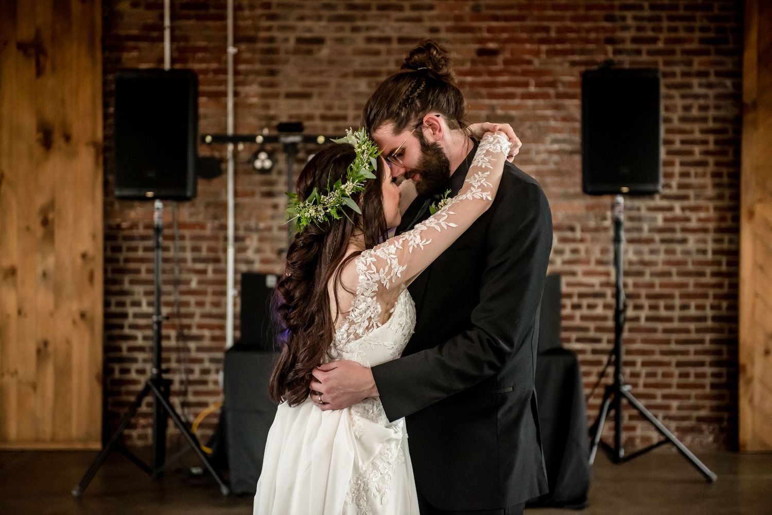 Bride and groom share first dance embracing in rustic brick venue with speakers and warm lighting.