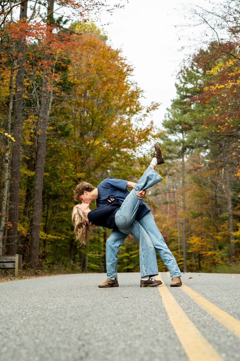 Playful couple poses on yellow road lines during fall photoshoot.