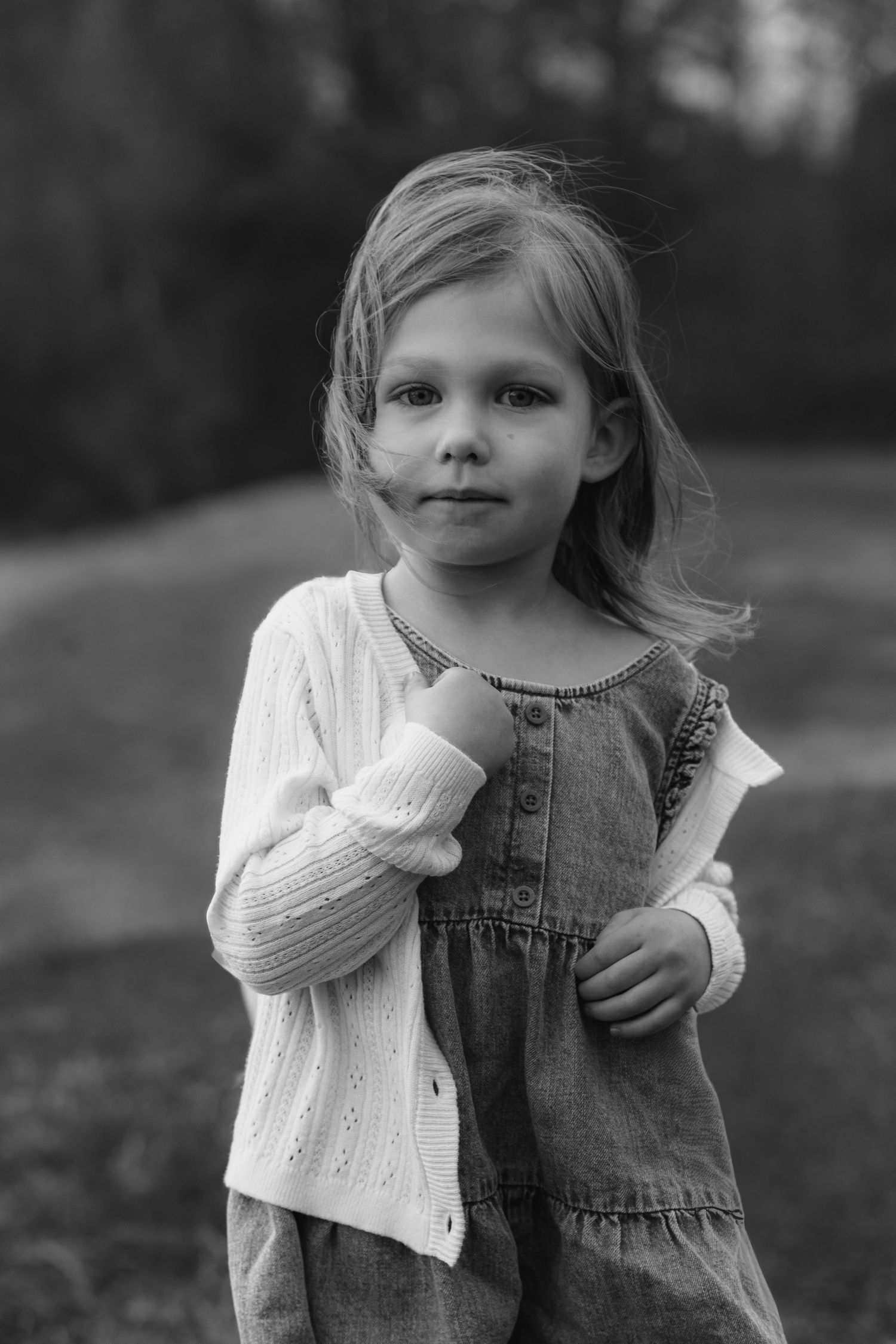 Black and white portrait of a young child wearing a denim dress and white cardigan sweater in an outdoor setting.