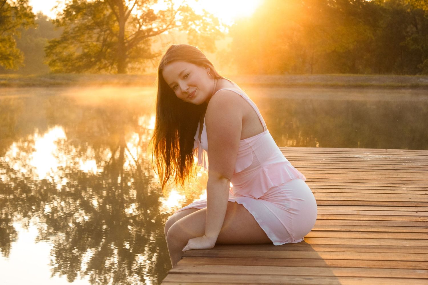 Person in white dress poses on wooden dock during golden sunset over tranquil lake.