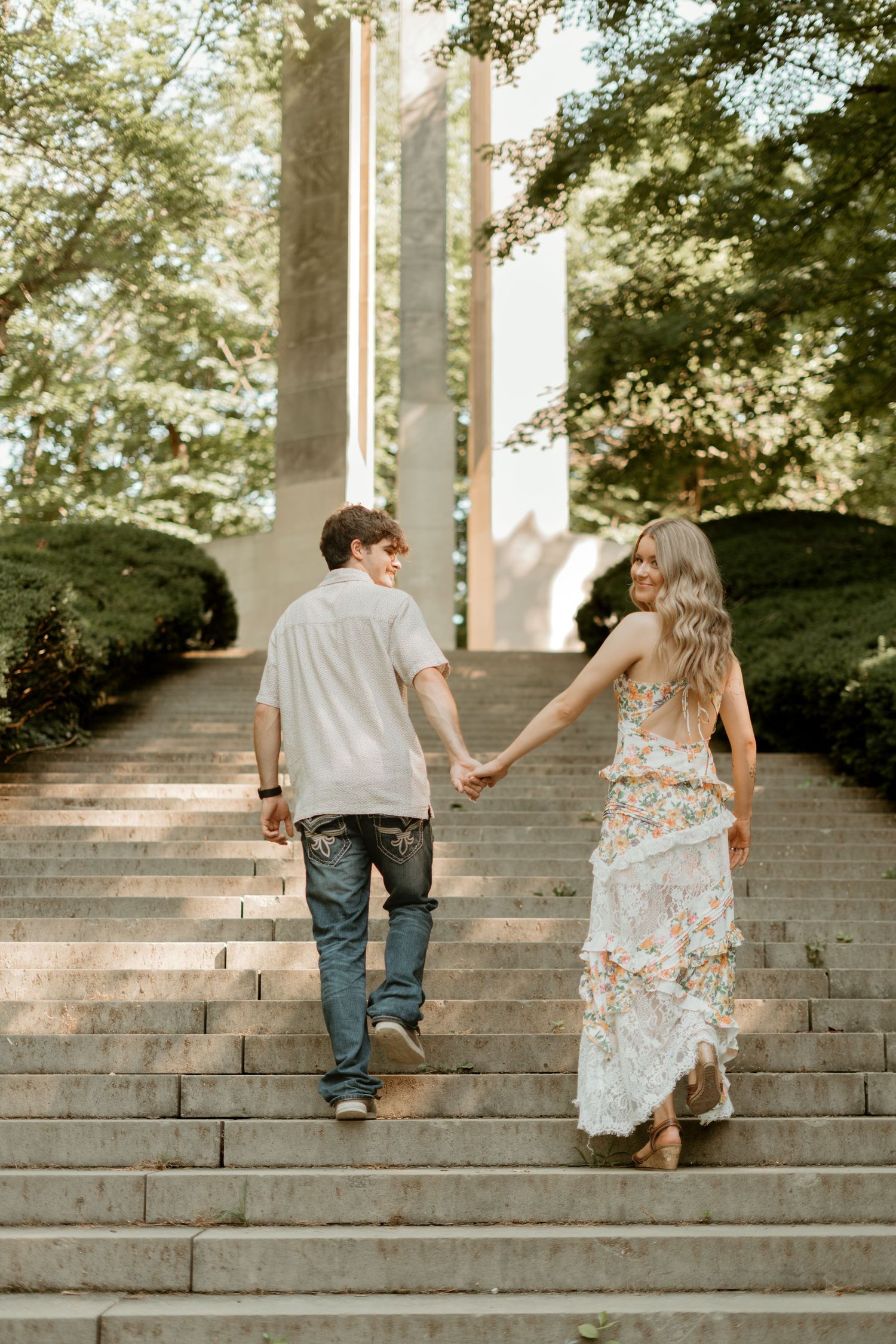 A couple holding hands while walking up stone steps together in a romantic outdoor setting with trees in the background.