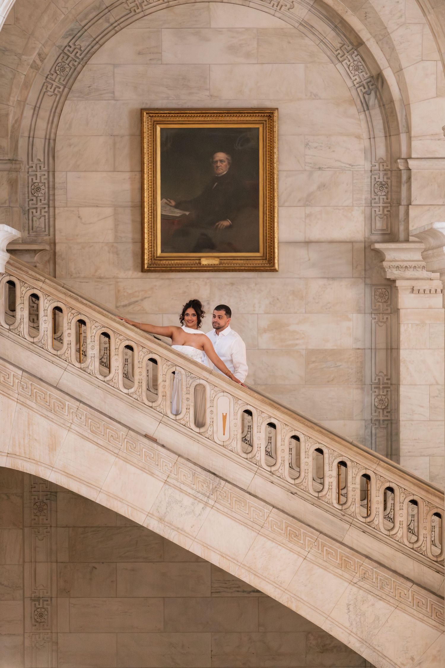 Bride and groom pose on grand marble staircase beneath portrait painting in elegant classical building.