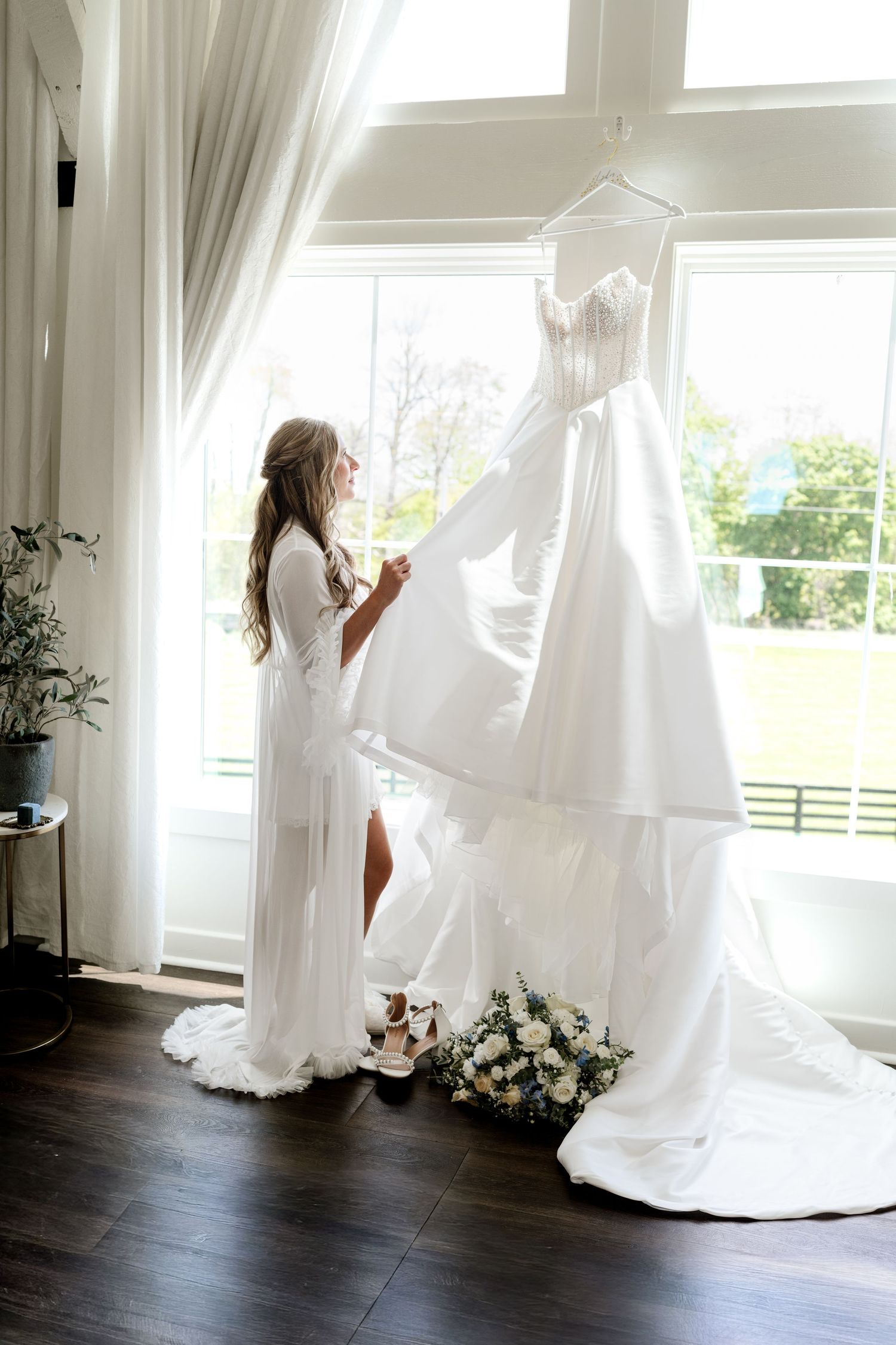 Bride in white robe admiring her elegant strapless wedding gown hanging by a bright window indoors.