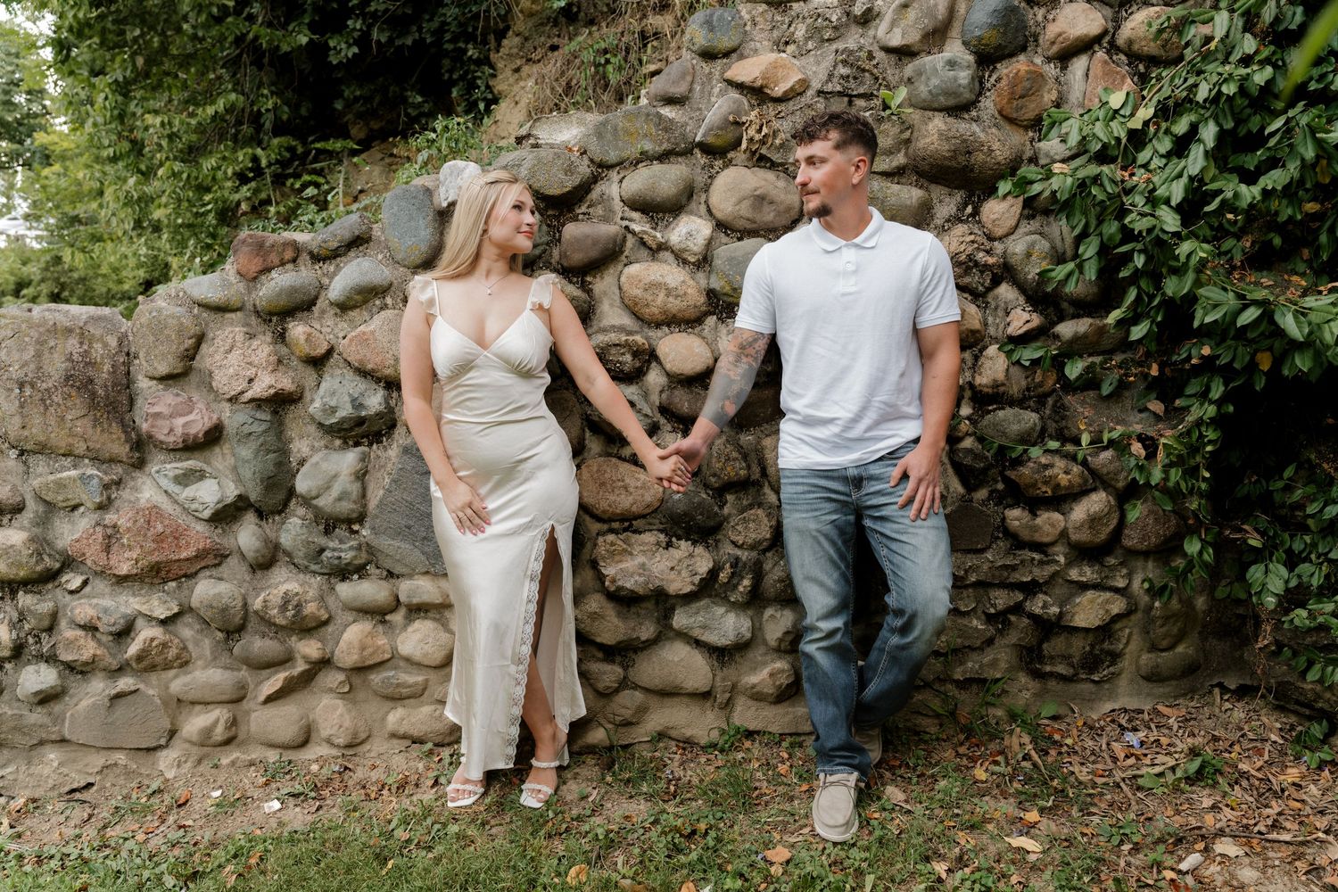 Couple posing against rustic stone wall in casual wedding attire.