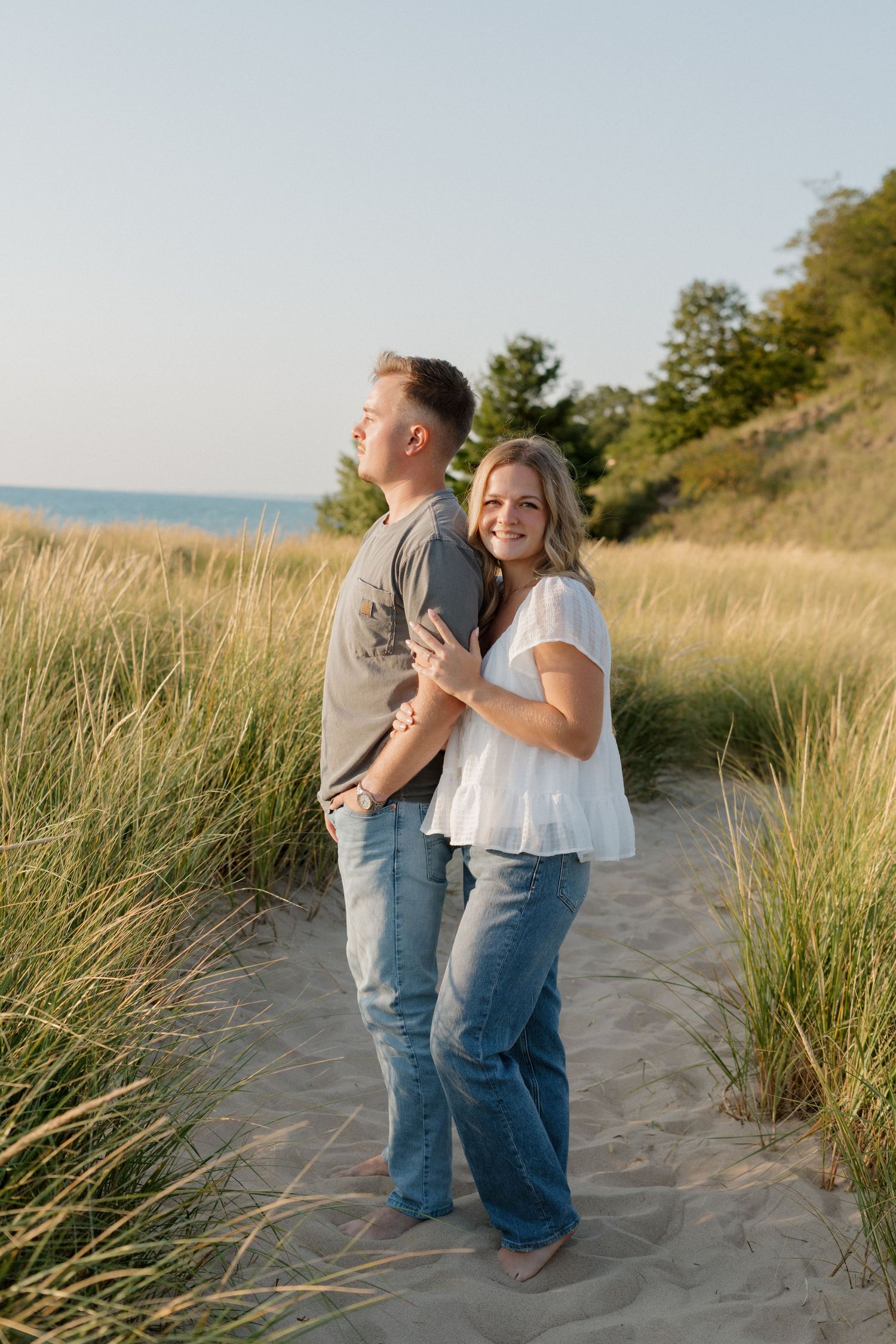 Romantic couple embracing on sandy beach trail surrounded by dune grass at sunset.