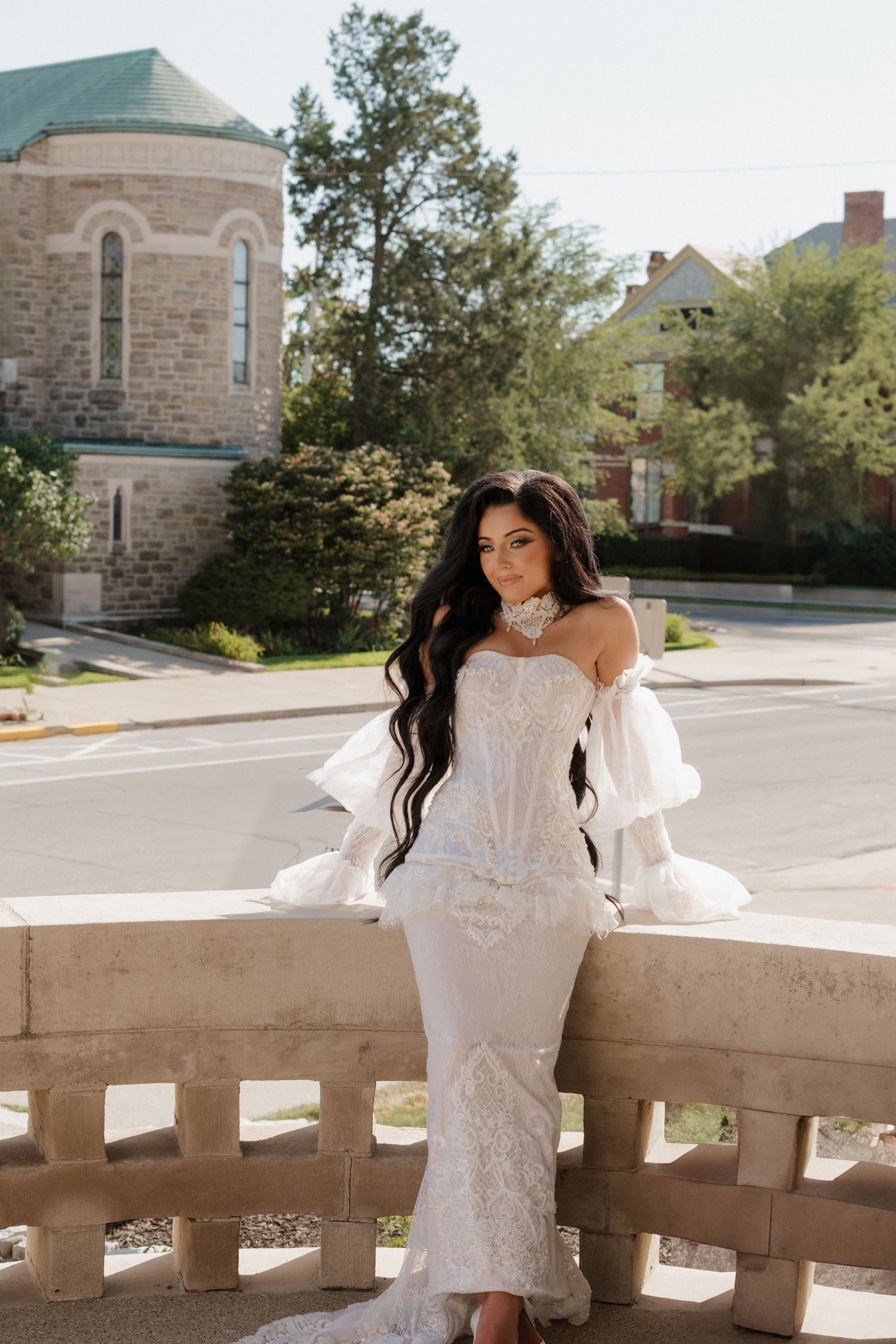 Bride in off-shoulder white gown poses along stone balustrade.