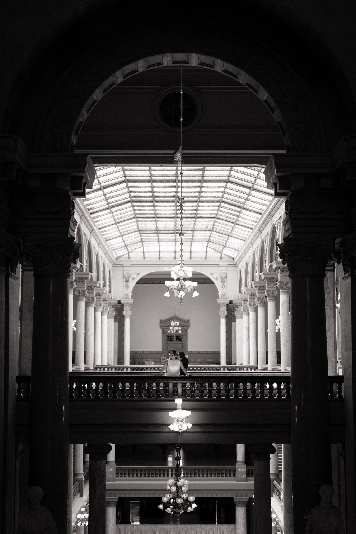 Black and white photo of a couple on a grand balcony inside an elegant historic building with chandeliers.