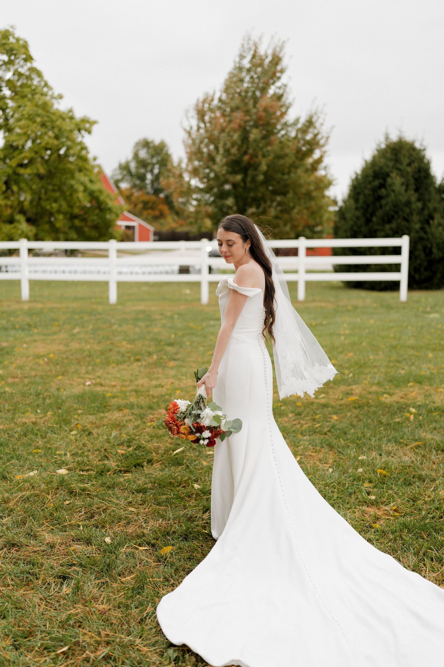 Bride in flowing white dress twirls gracefully on grass field with white fence in background.
