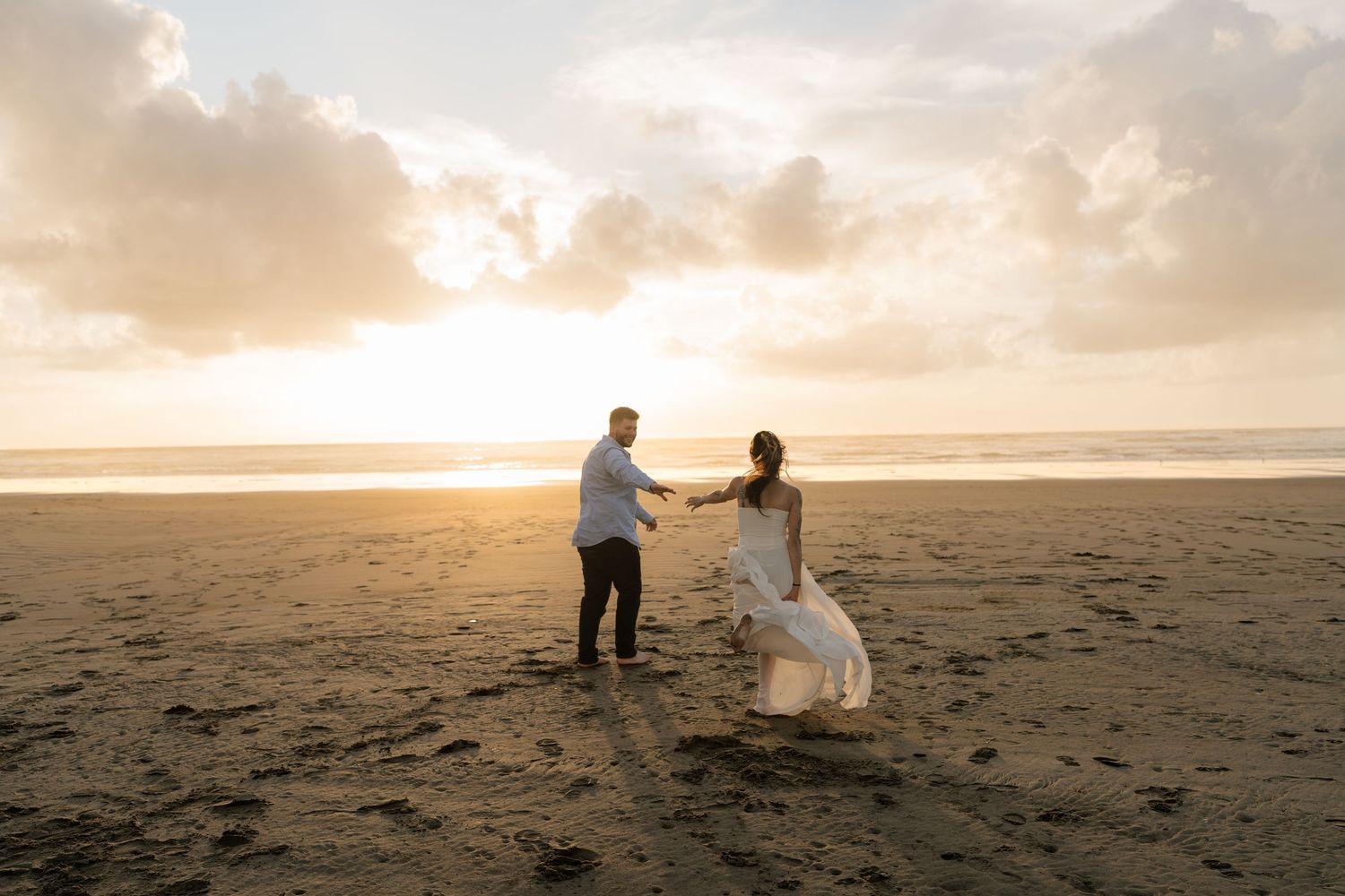 Bride and groom hold hands on a sandy beach at golden sunset during romantic wedding photo session.