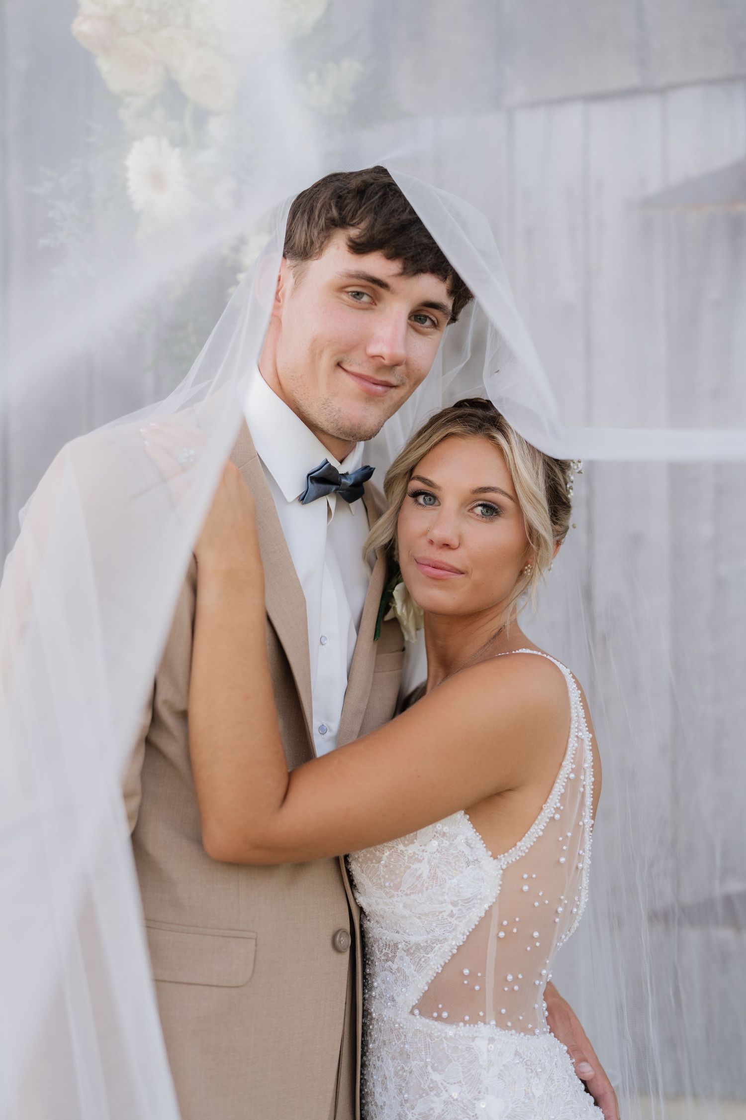 Bride and groom posing under a flowing white veil, groom in tan suit with bow tie, bride in beaded wedding dress.