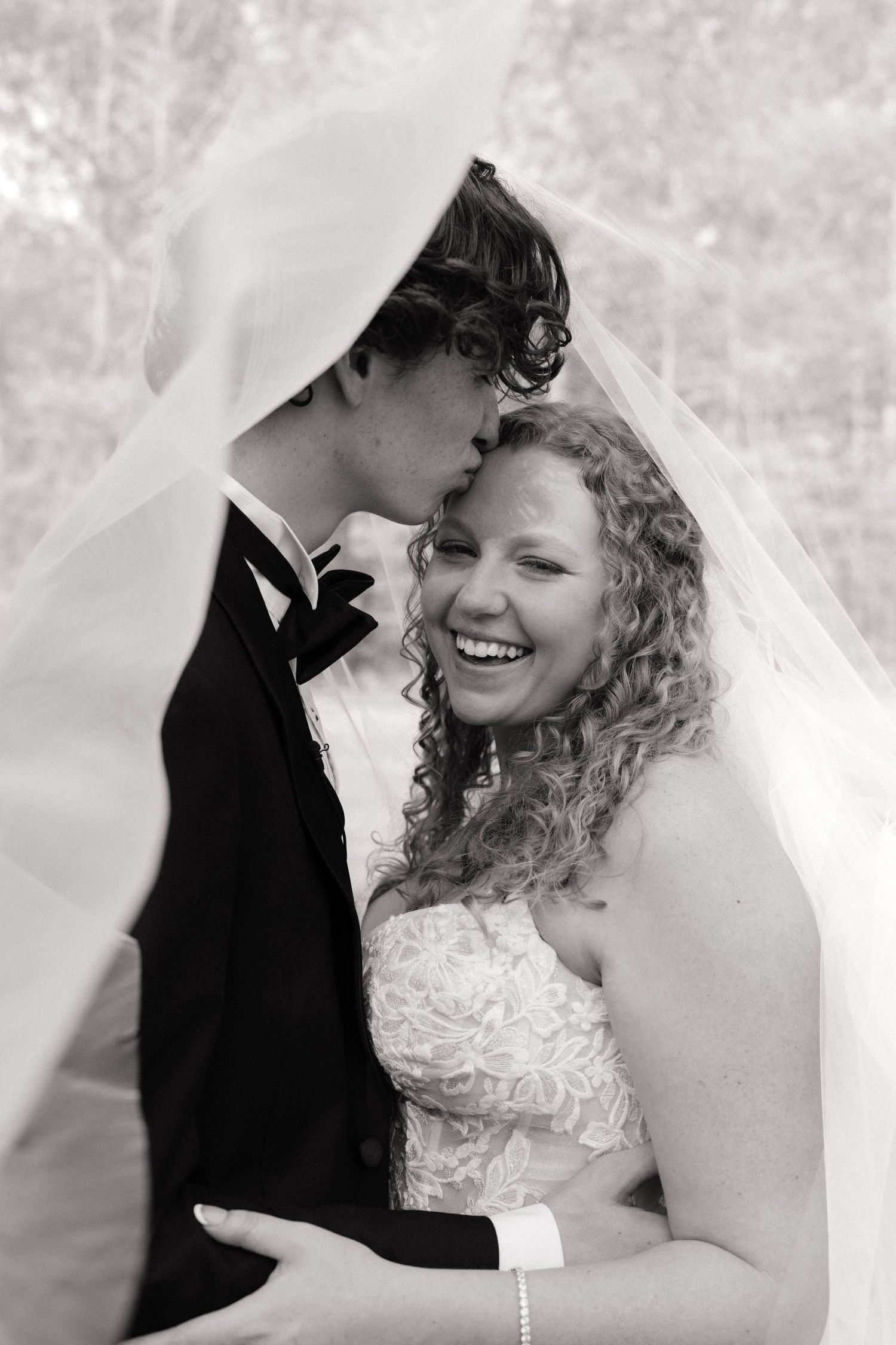 Black and white photo of a smiling bride with curly hair and groom kissing her cheek under a flowing veil.
